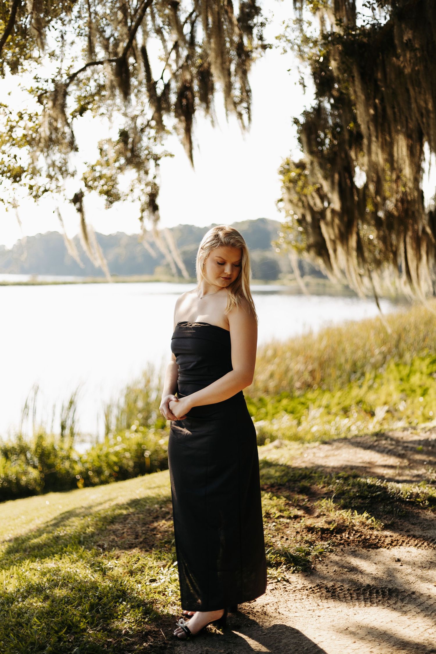 A person in a black evening dress stands along a scenic waterfront path beneath moss-draped trees at sunset.