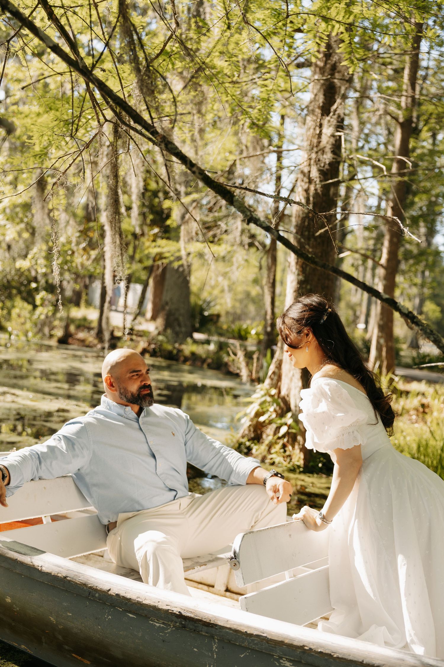 A couple in white attire shares a romantic moment in a rowboat surrounded by lush trees near a tranquil lake.