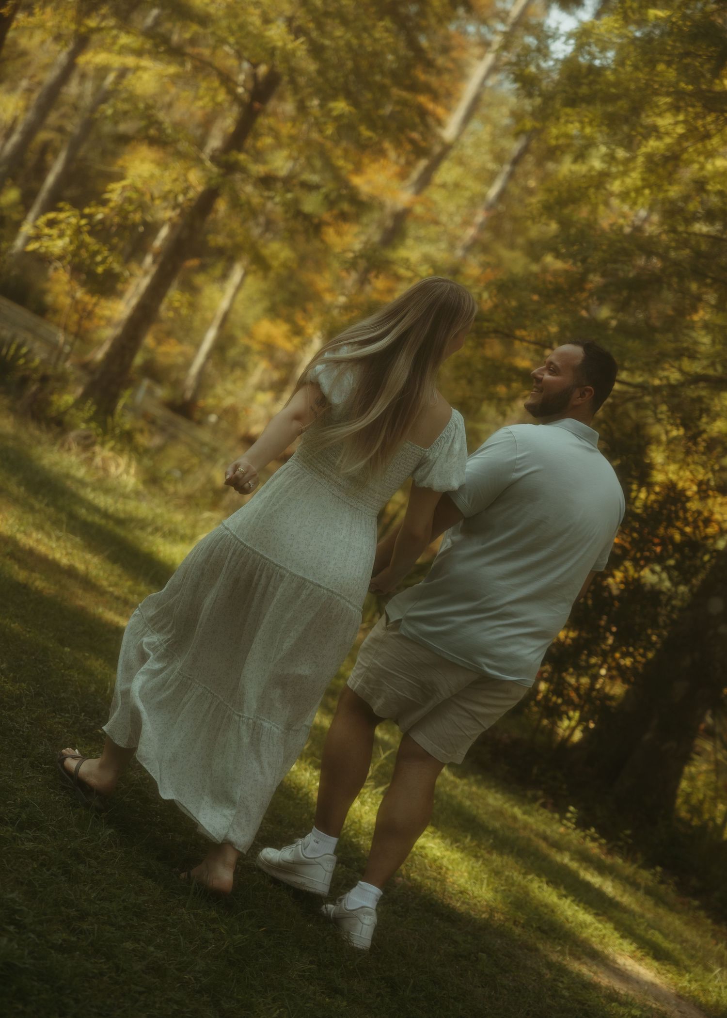 Couple holding hands and laughing in a lush autumn forest, woman in white dress, man in light blue shirt and shorts.