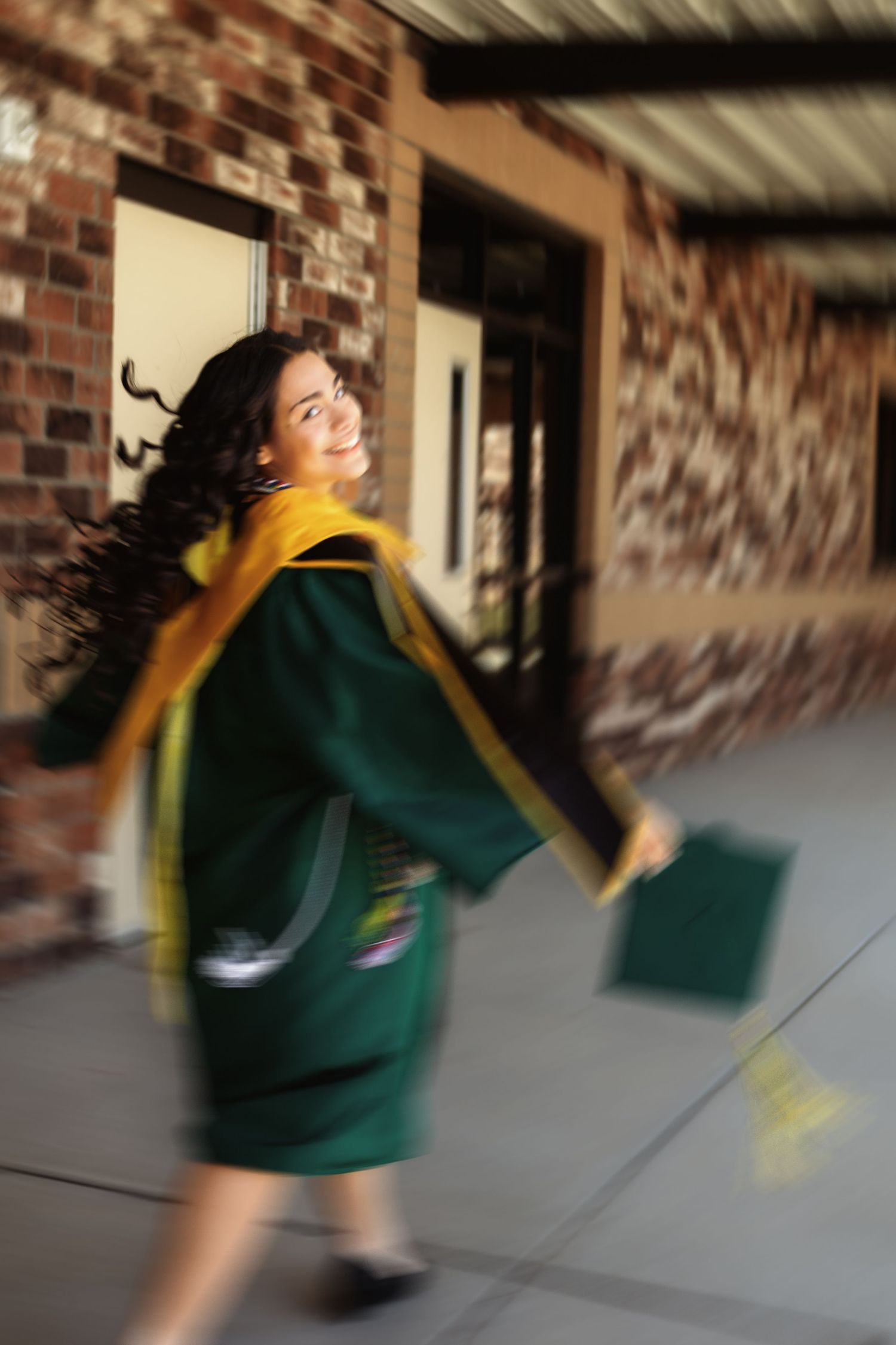 Motion blur captures graduate spinning in green gown along brick walkway.