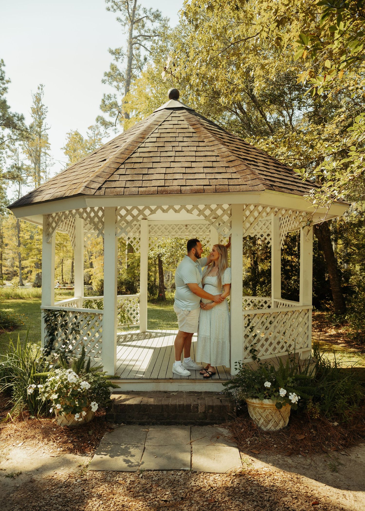 A couple shares a romantic moment inside a white lattice gazebo surrounded by lush green trees and flowers.