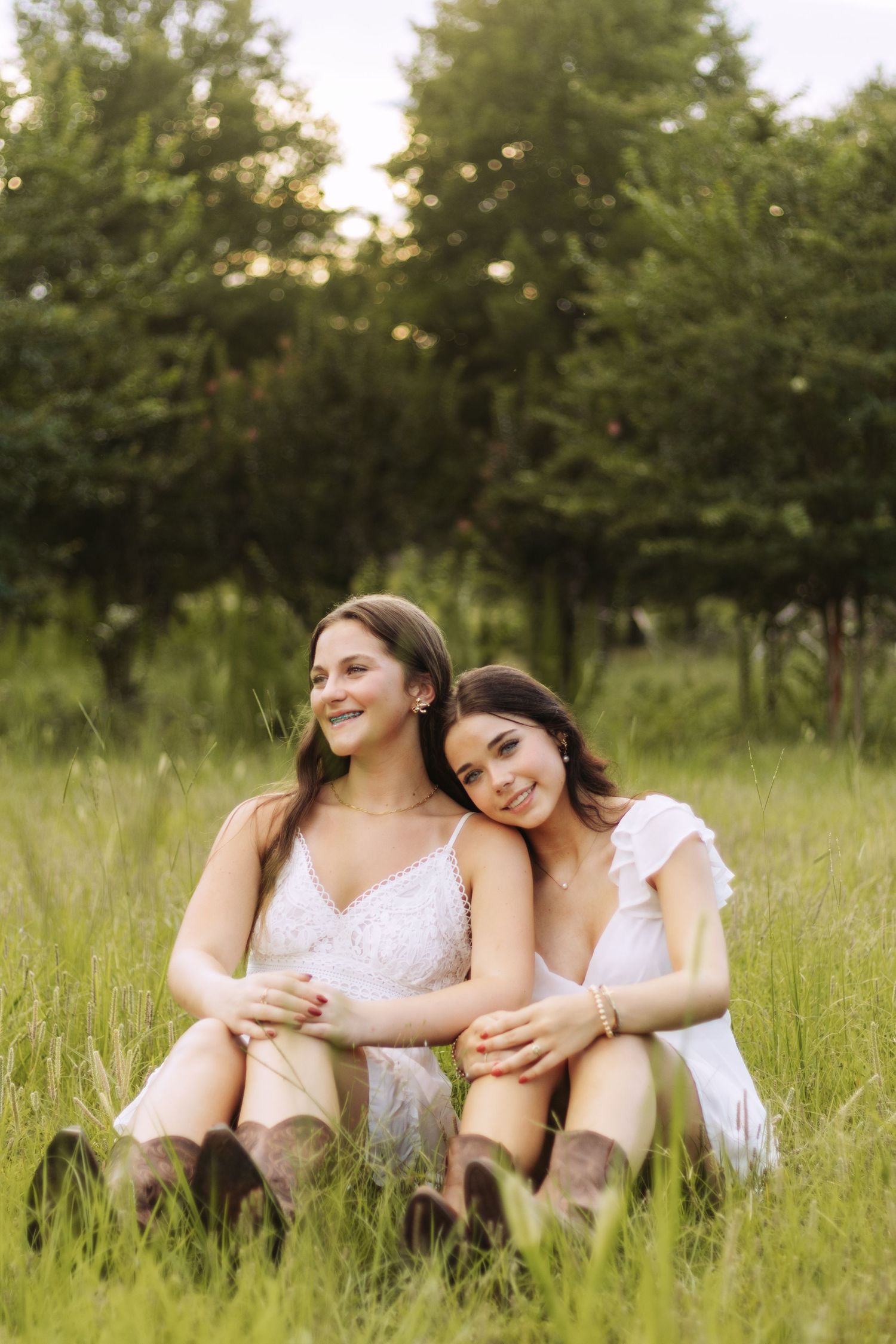 Two people in white dresses sit together laughing in a sunlit meadow with tall grass and trees in the background.