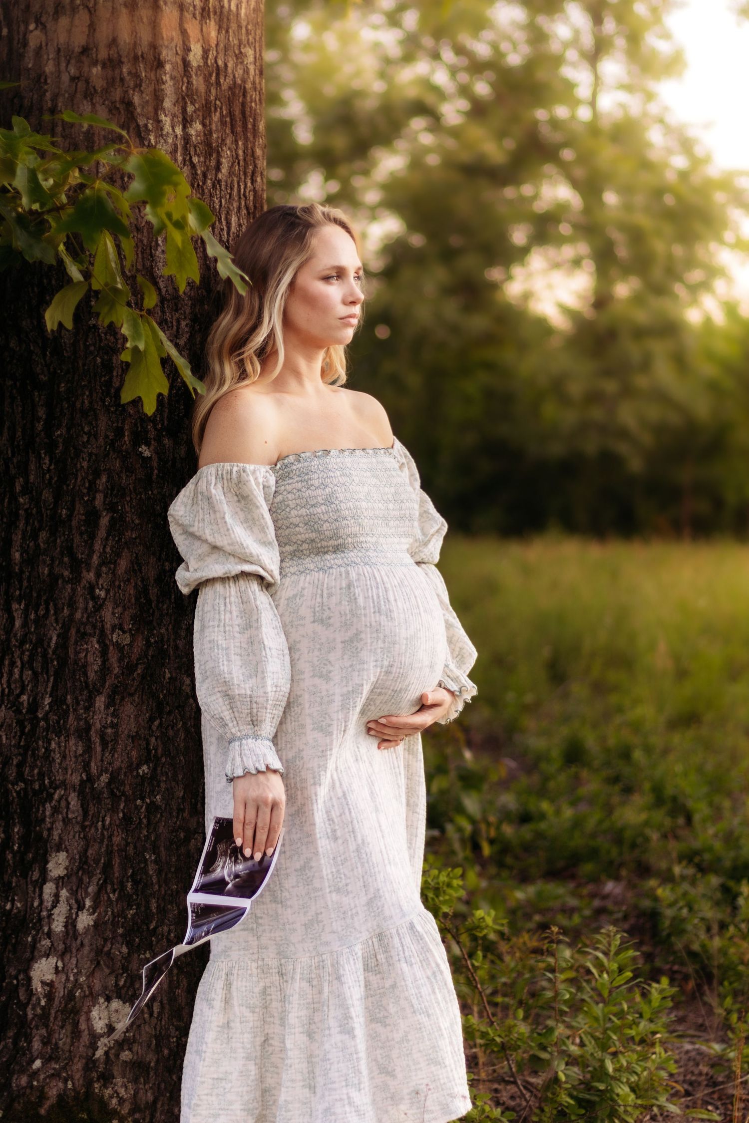 Person in white off-shoulder dress poses against tree trunk holding ultrasound photos.