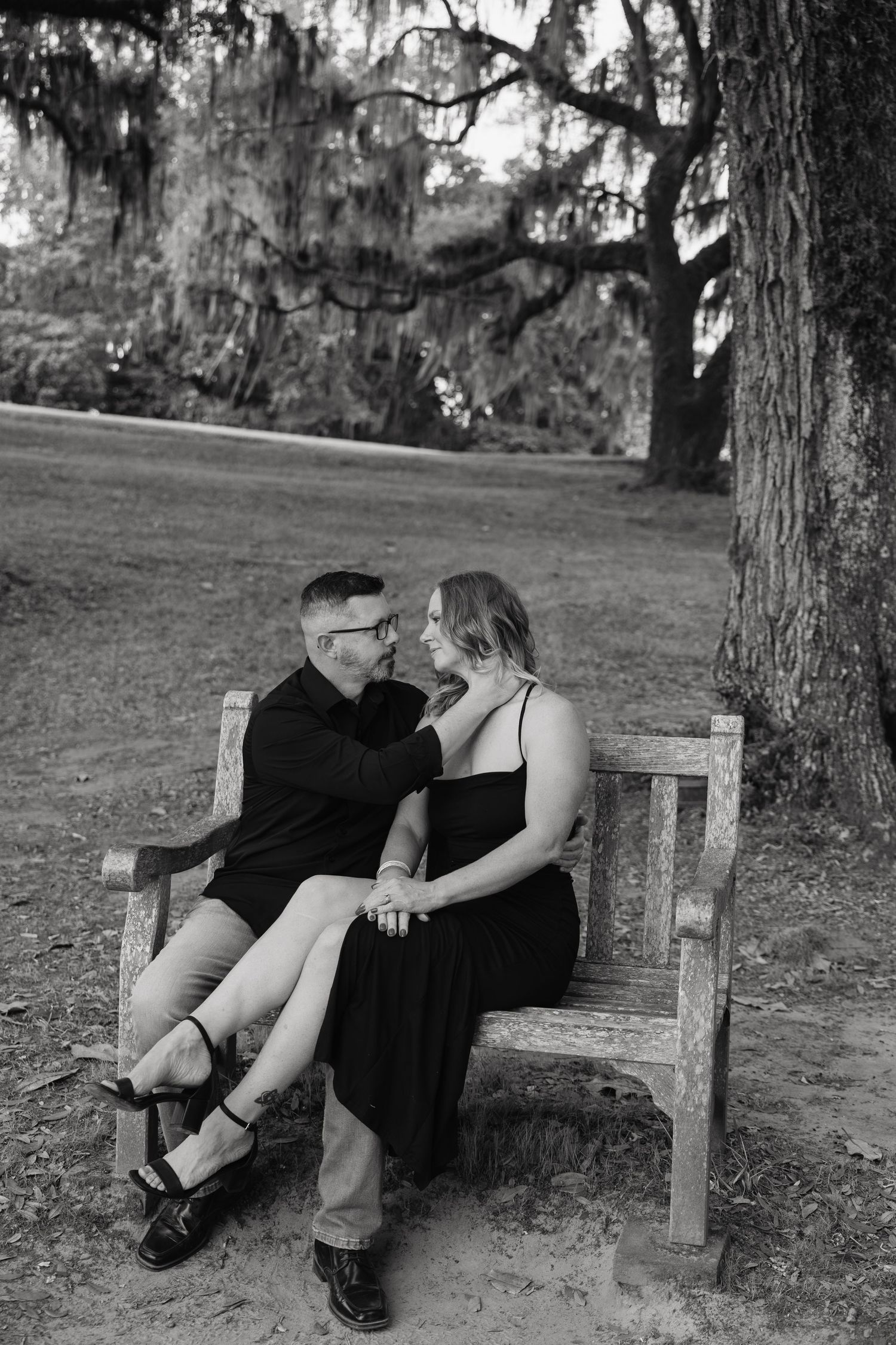 Couple sitting on a wooden bench under a moss-draped tree, holding hands and gazing at each other in black and white.