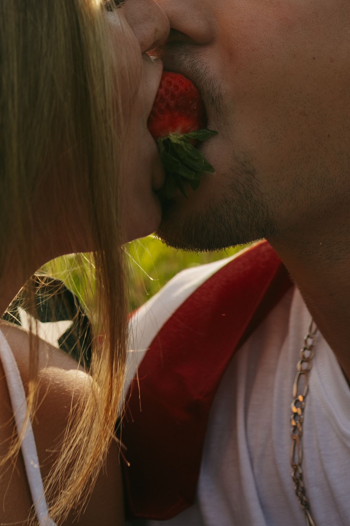 Couple sharing a fresh strawberry in a romantic kiss, with golden sunlight and lush green background.