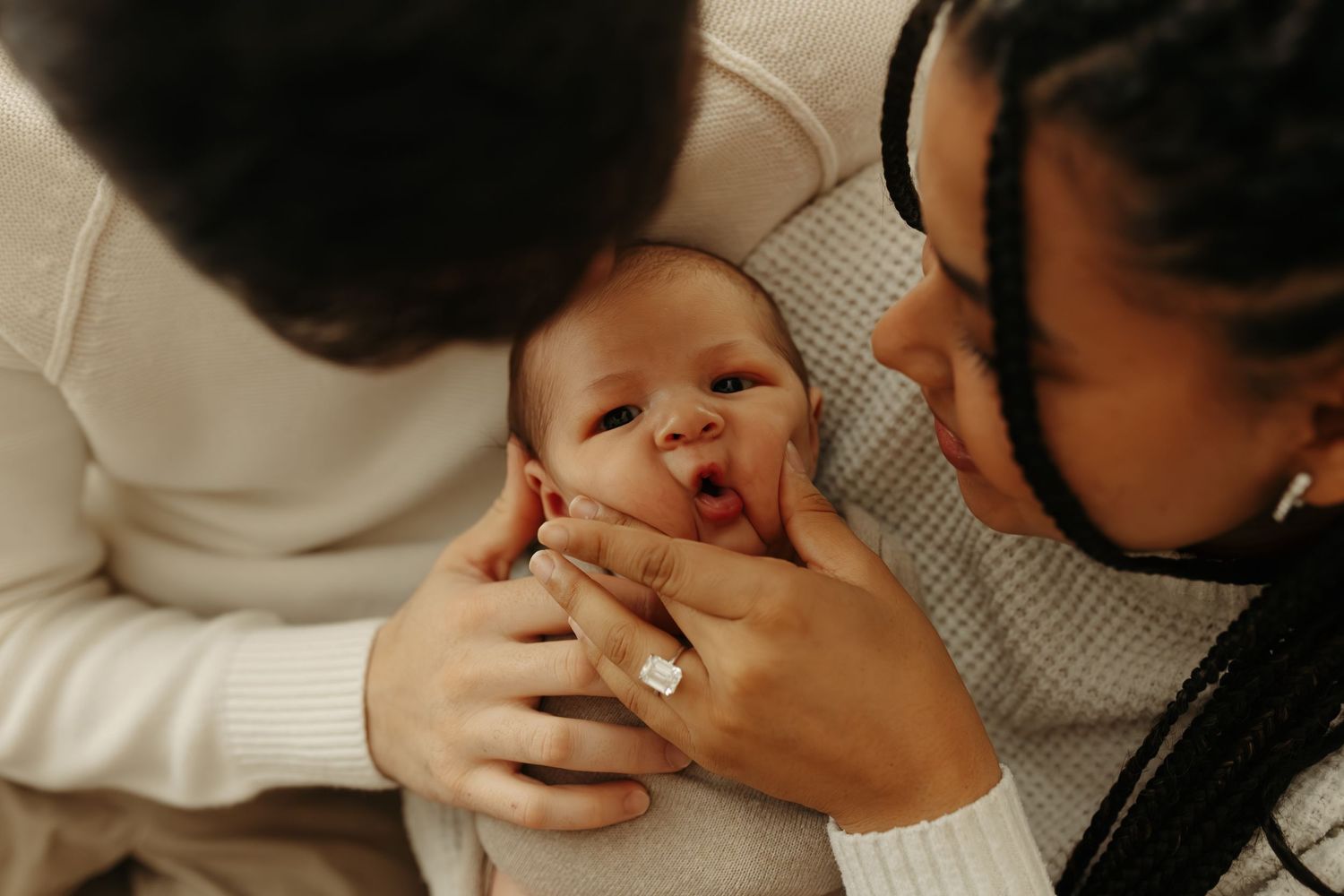 Two parents lovingly holding and kissing their newborn baby, dressed in white outfits.