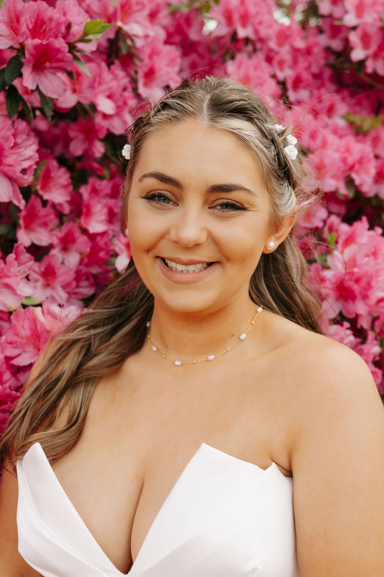 Young woman in white dress with pearl necklace and hair accessories smiling in front of pink azalea flowers.