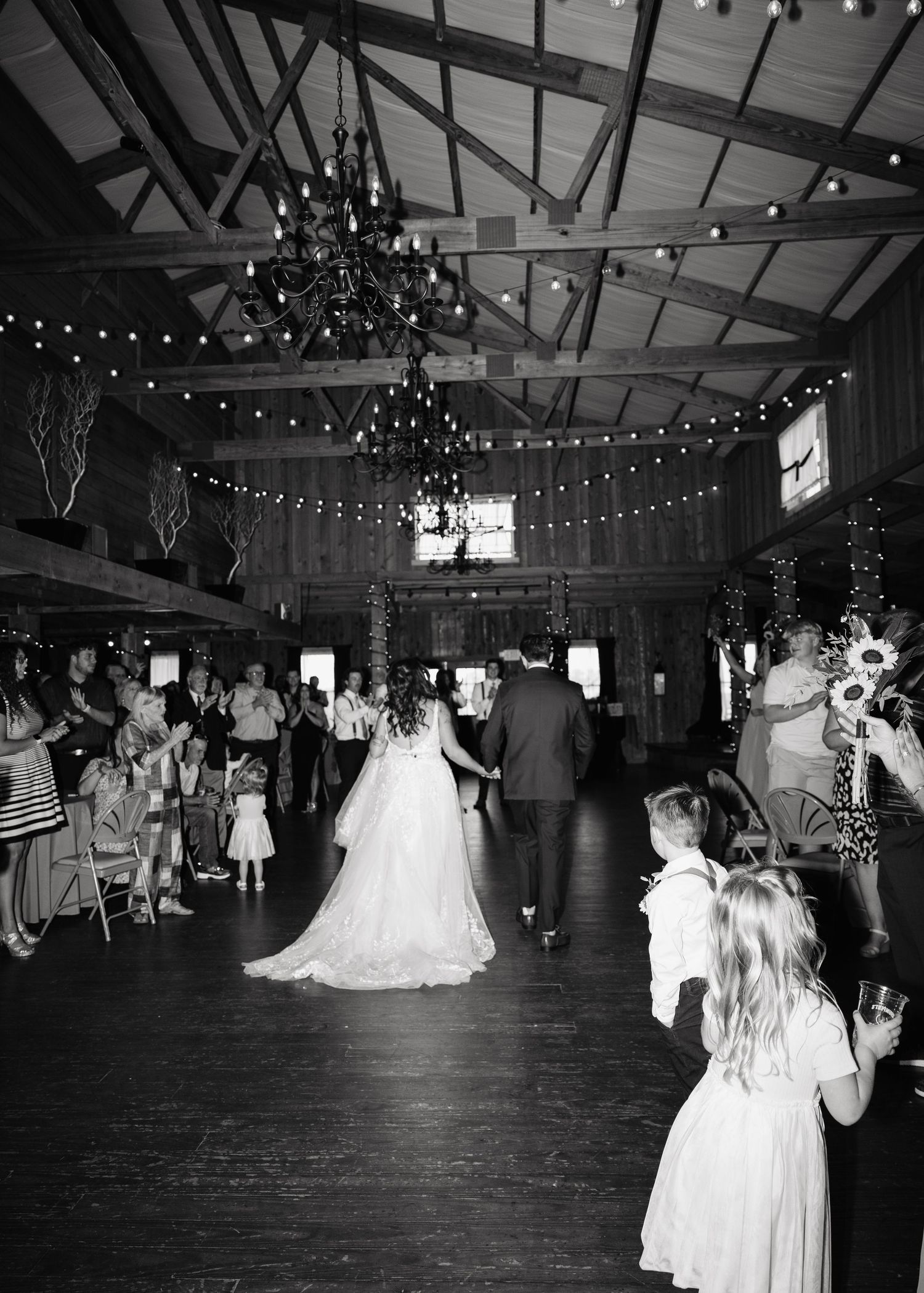 Bride and groom make grand entrance into rustic barn reception with string lights overhead.