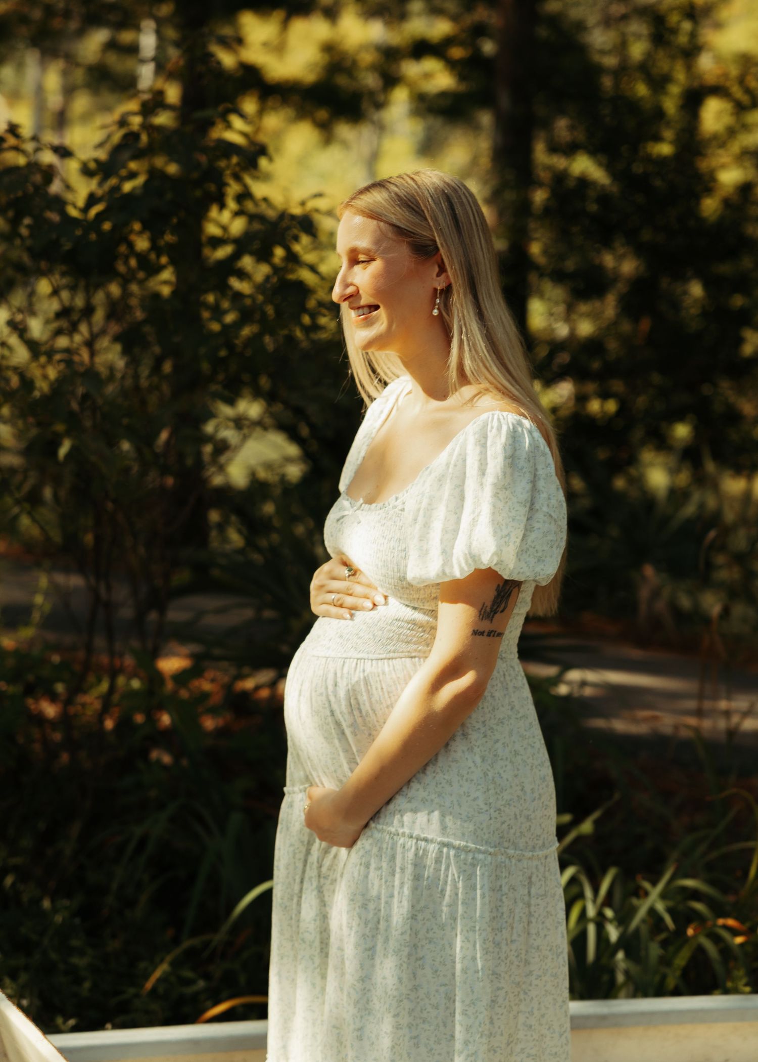 Smiling pregnant woman in white floral dress cradling baby bump outdoors in golden sunlight among lush green trees.