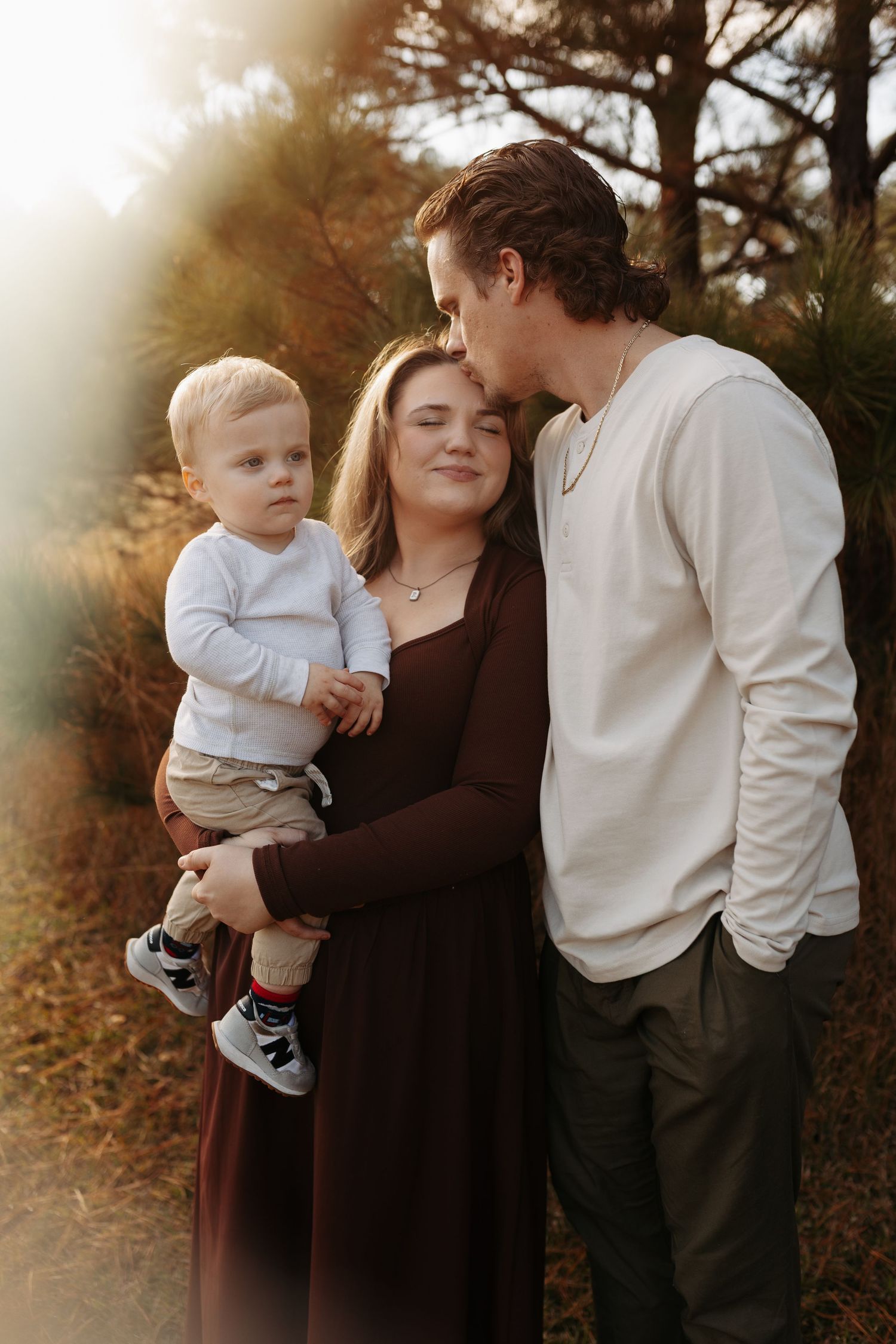 A young family poses outdoors, mother holding toddler while father kisses her cheek, surrounded by warm autumn foliage.