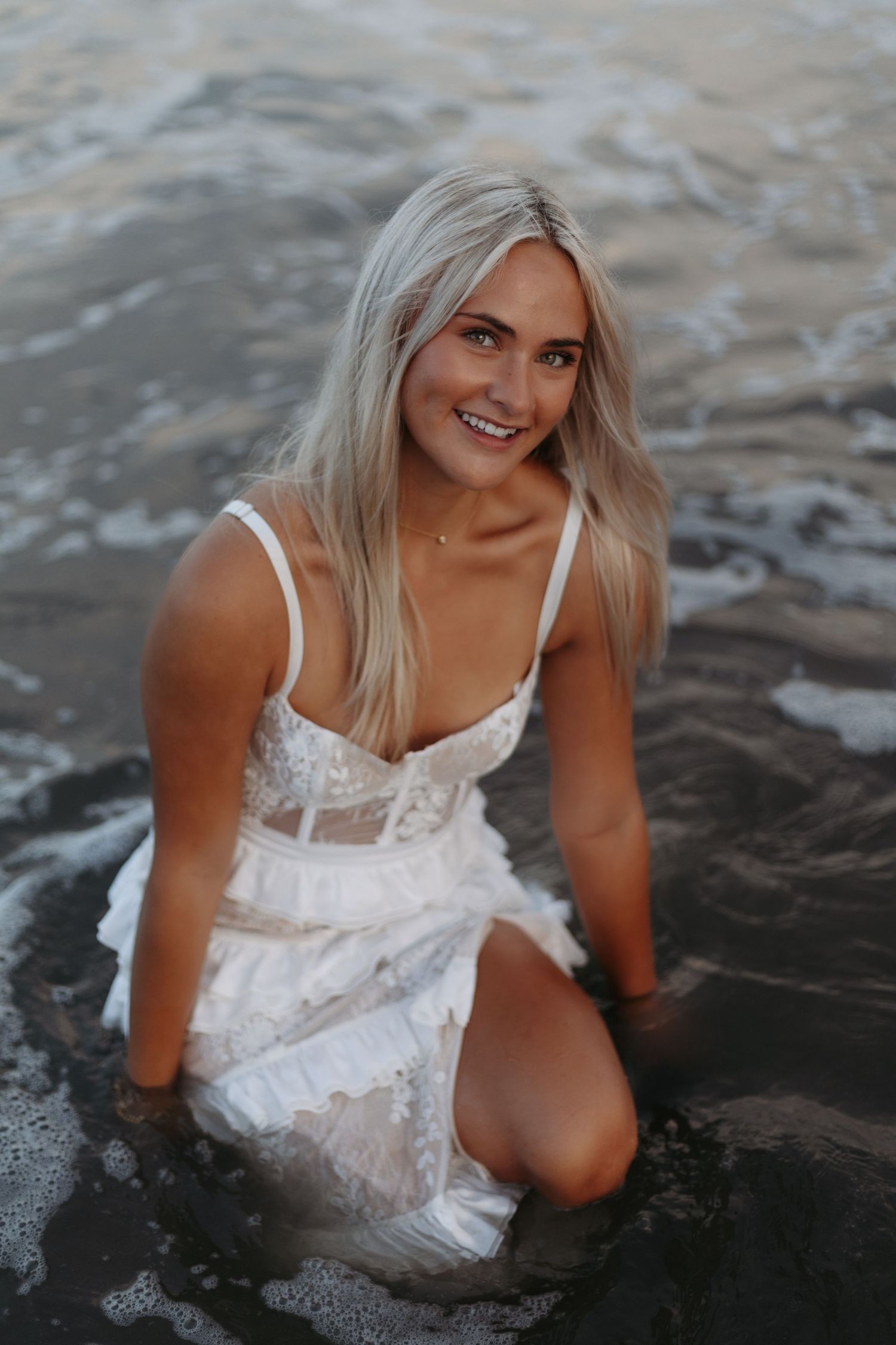 Smiling blonde woman in white lace dress sitting in ocean waves on a sandy beach at golden hour.