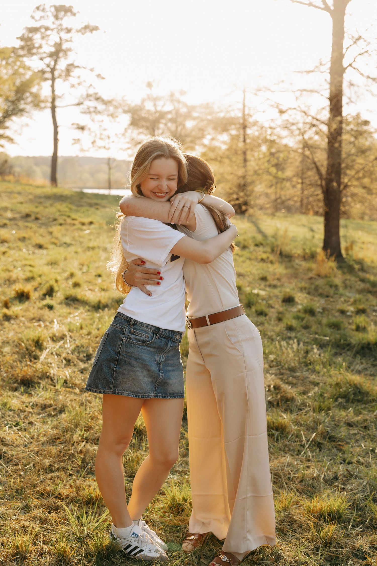 Two young women share a warm hug outdoors in golden sunset light, standing on green grass with trees behind them.