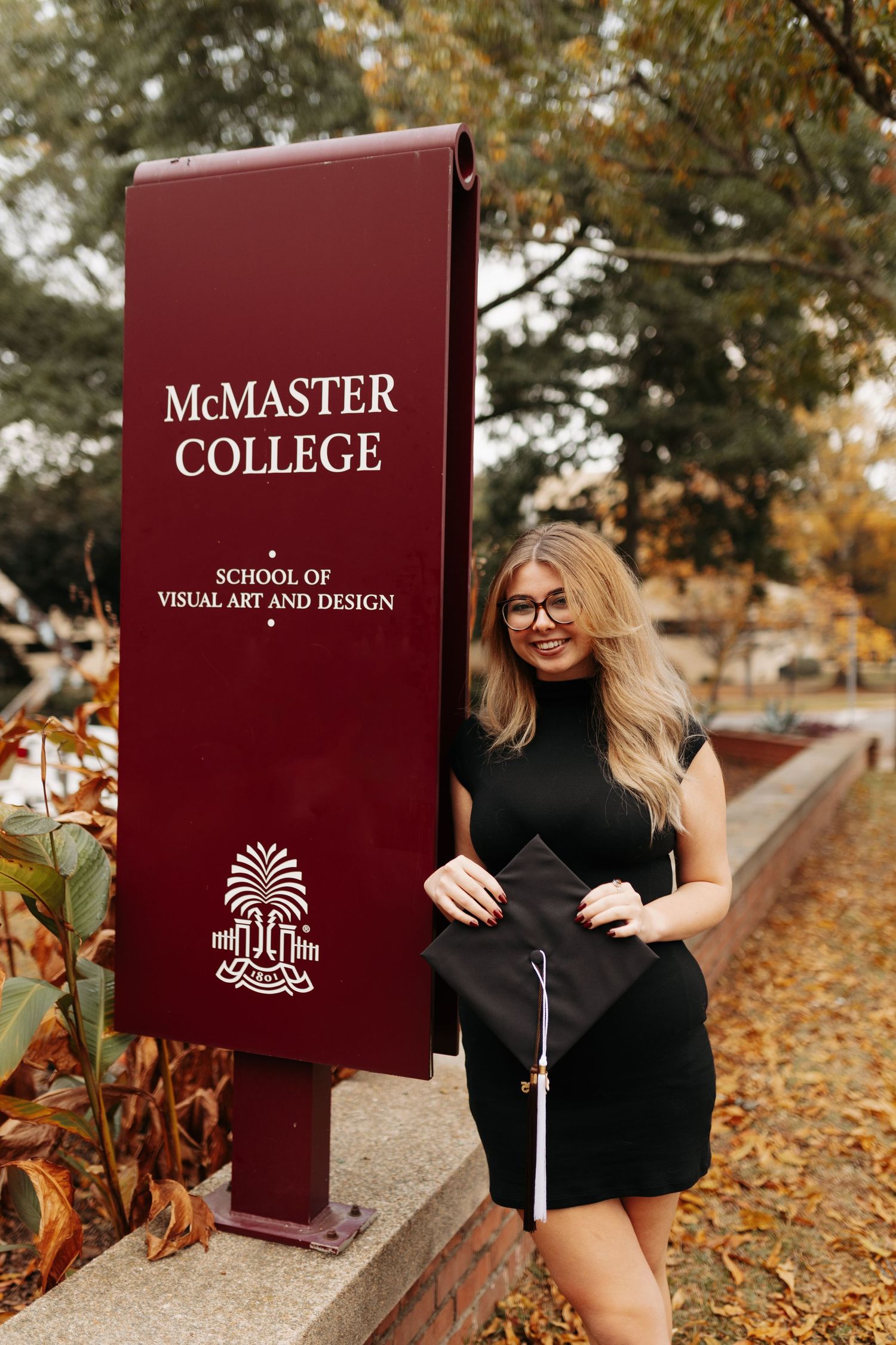 A smiling graduate in a black dress holds her cap beside a McMaster College School of Visual Art and Design sign.