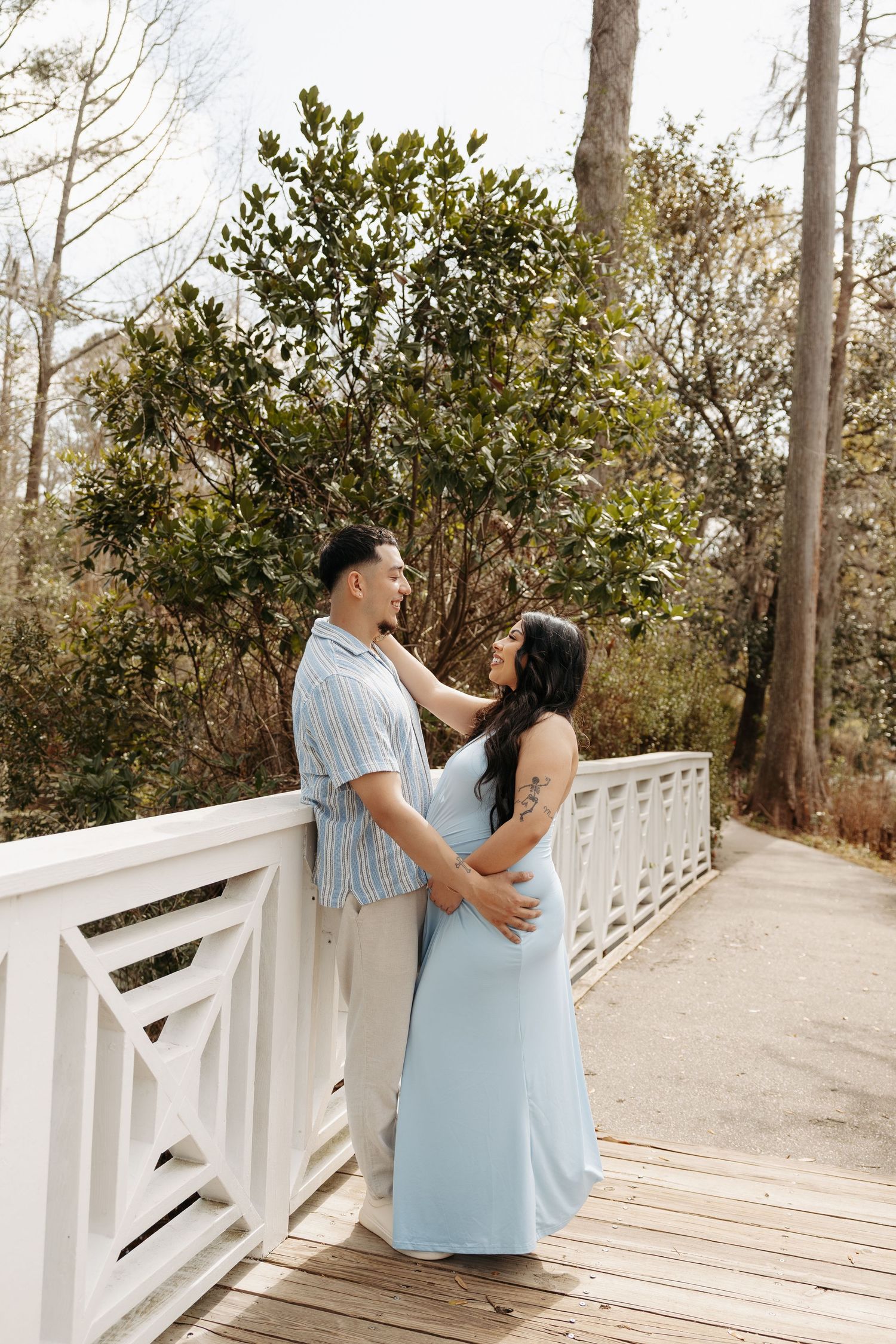 A couple in matching light blue outfits share a romantic moment on a white wooden bridge surrounded by lush trees.