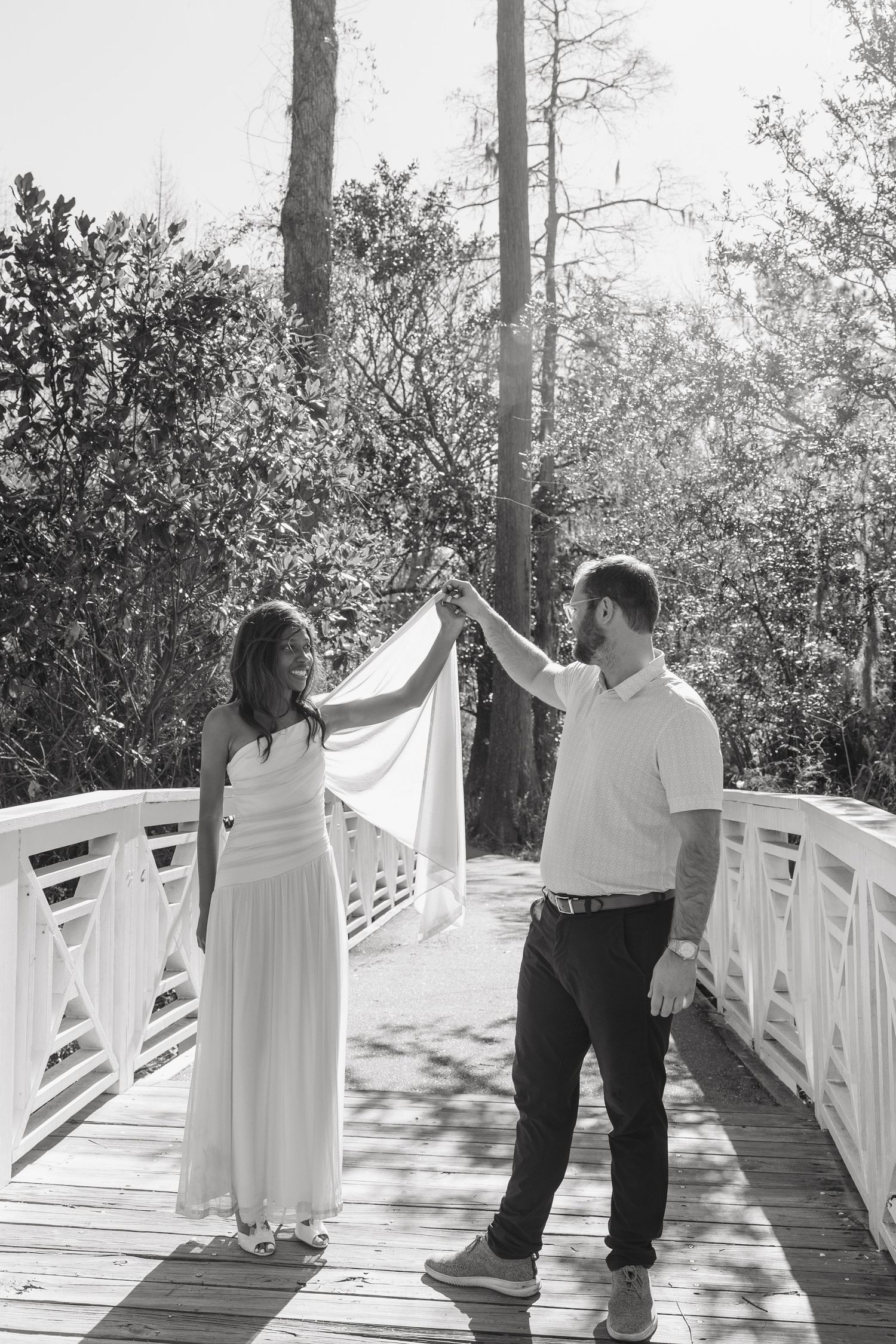 Black and white photo of a couple on a wooden bridge, man lifting woman's white dress veil outdoors.