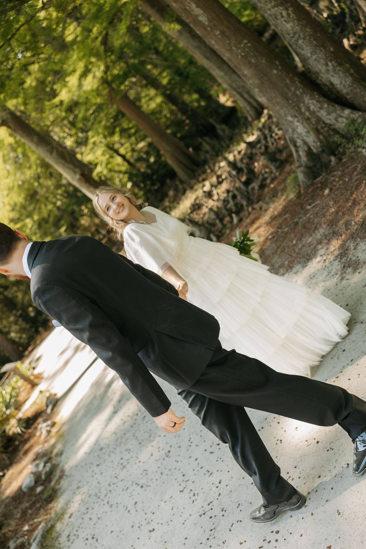 A smiling bride in a white gown holds hands with a groom in a black suit while walking through a forest path.
