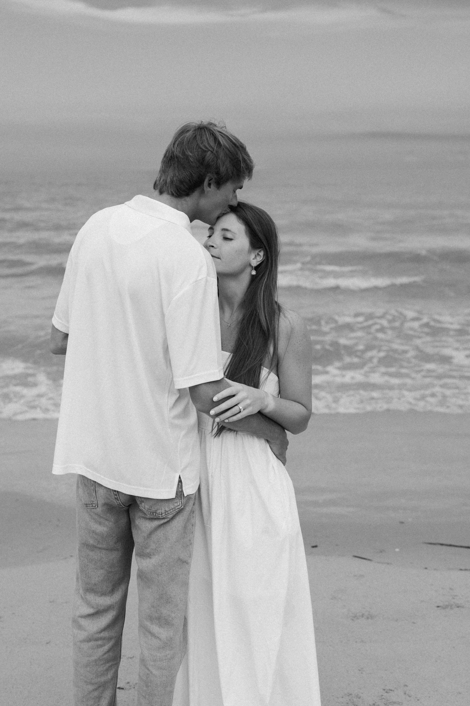 Black and white photo of a romantic couple embracing on a beach, woman in white dress, waves in background.