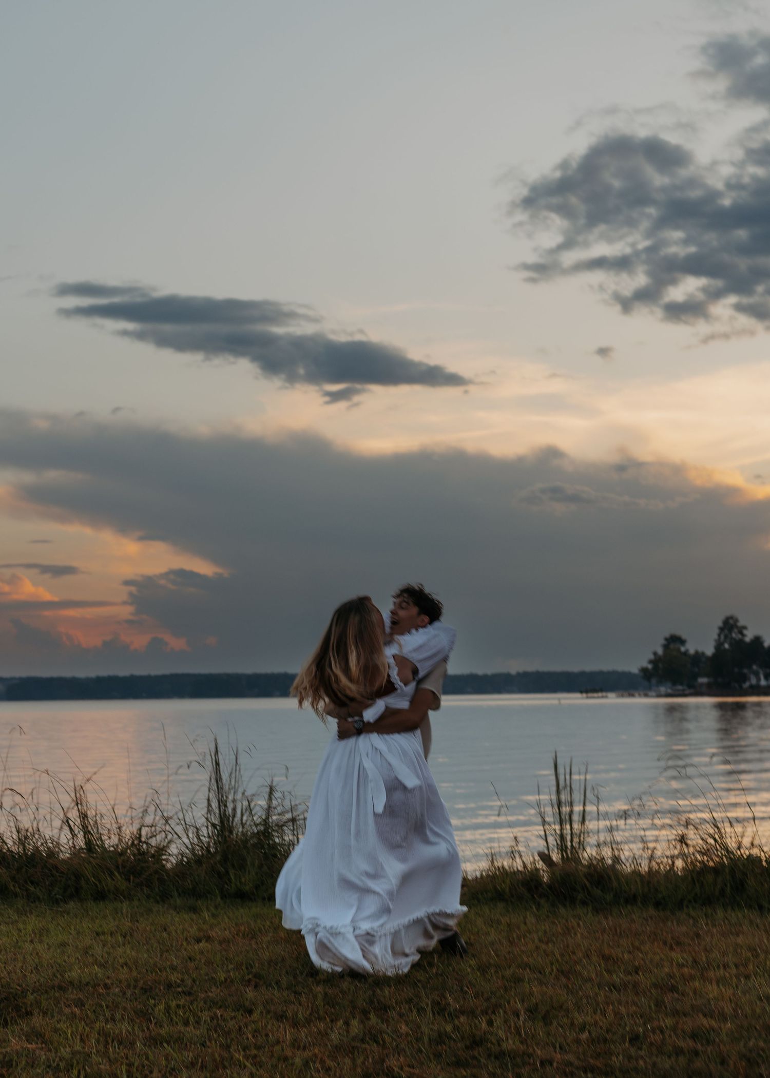 A mother in a white dress holds her baby by a serene lakeside at sunset, sharing a tender moment outdoors.