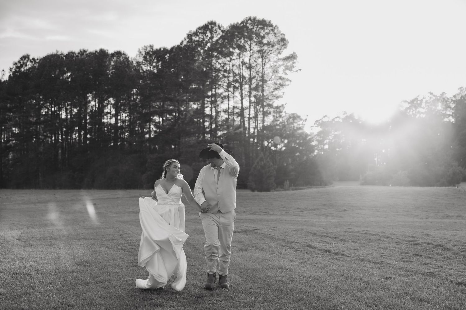 Black and white photo of a bride and groom holding hands in a sunlit field with tall pine trees in the background.