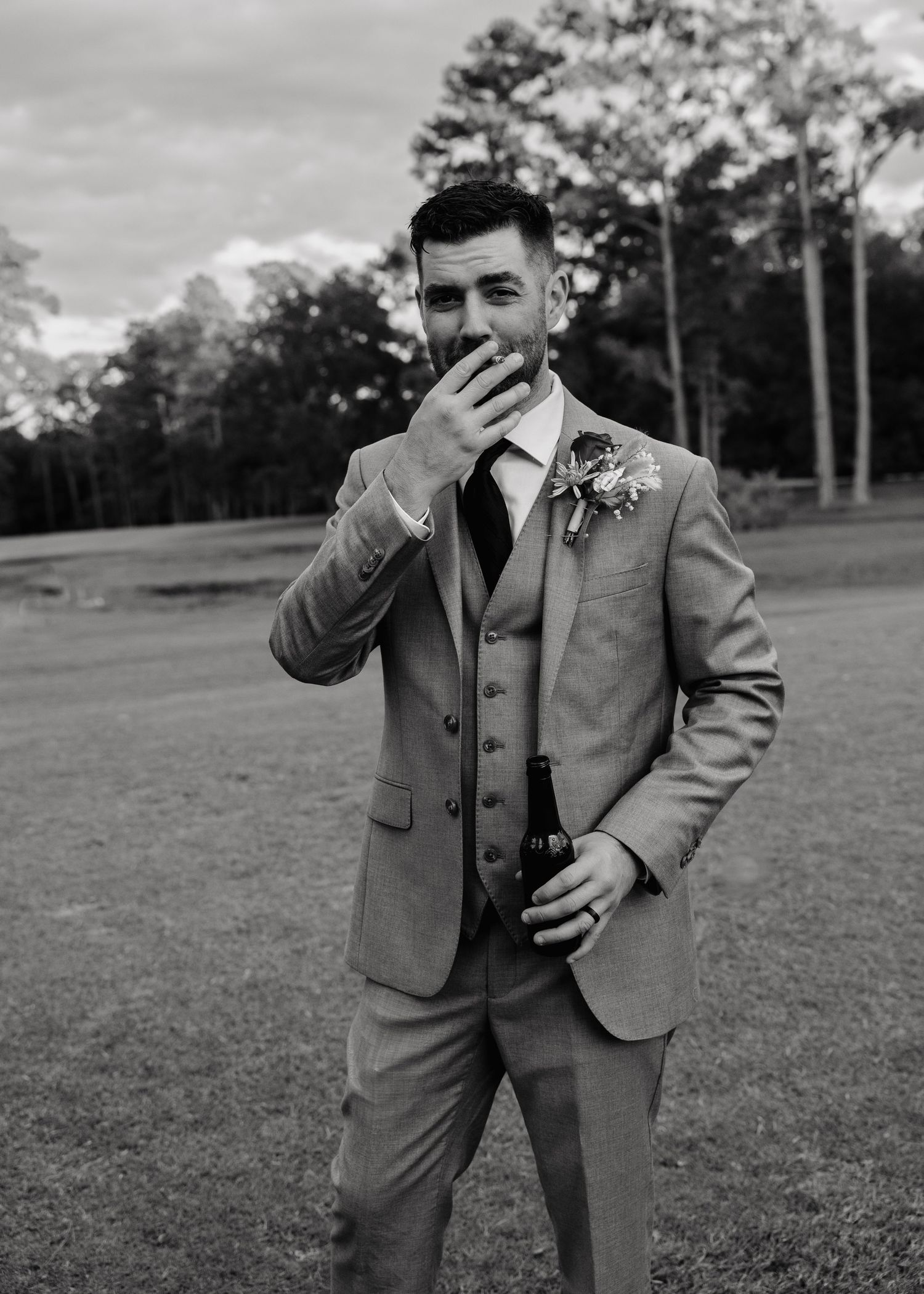 Dapper groom in a gray three-piece suit smoking a cigar while holding a champagne bottle outdoors.