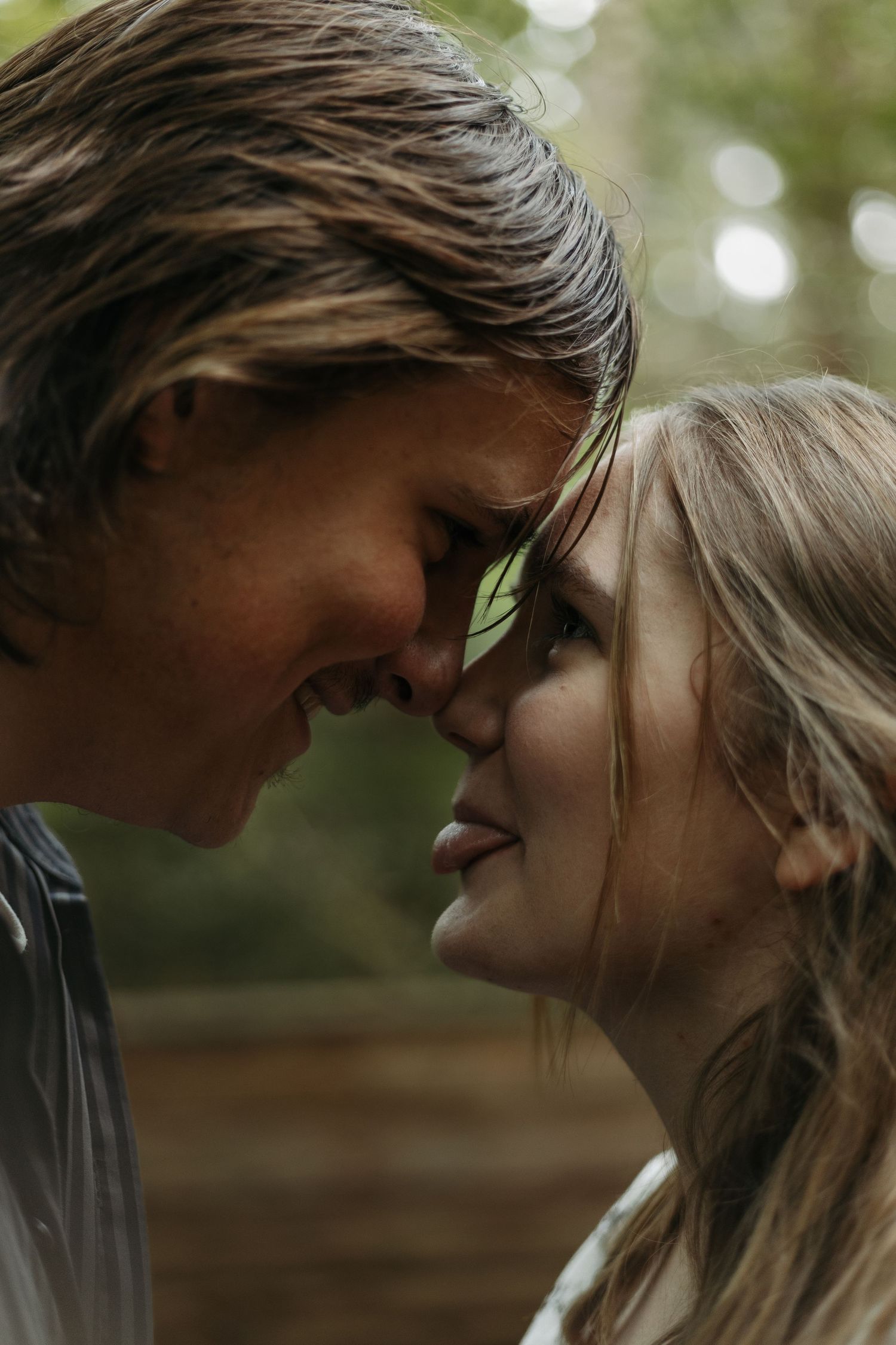 A young couple sharing a tender moment, foreheads touching and smiling in a lush green outdoor setting.
