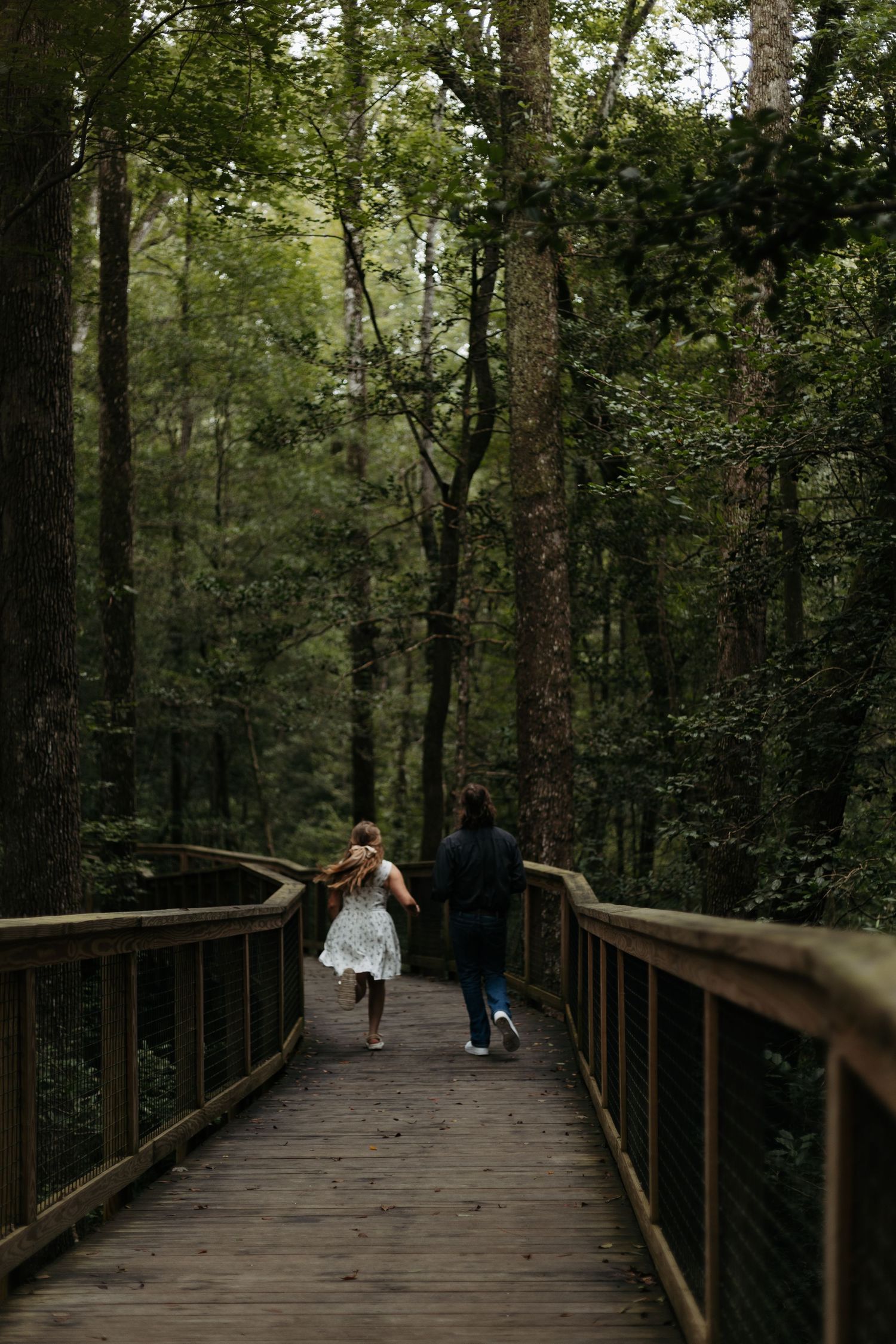 Two people walk along a wooden boardwalk trail surrounded by lush green forest trees.