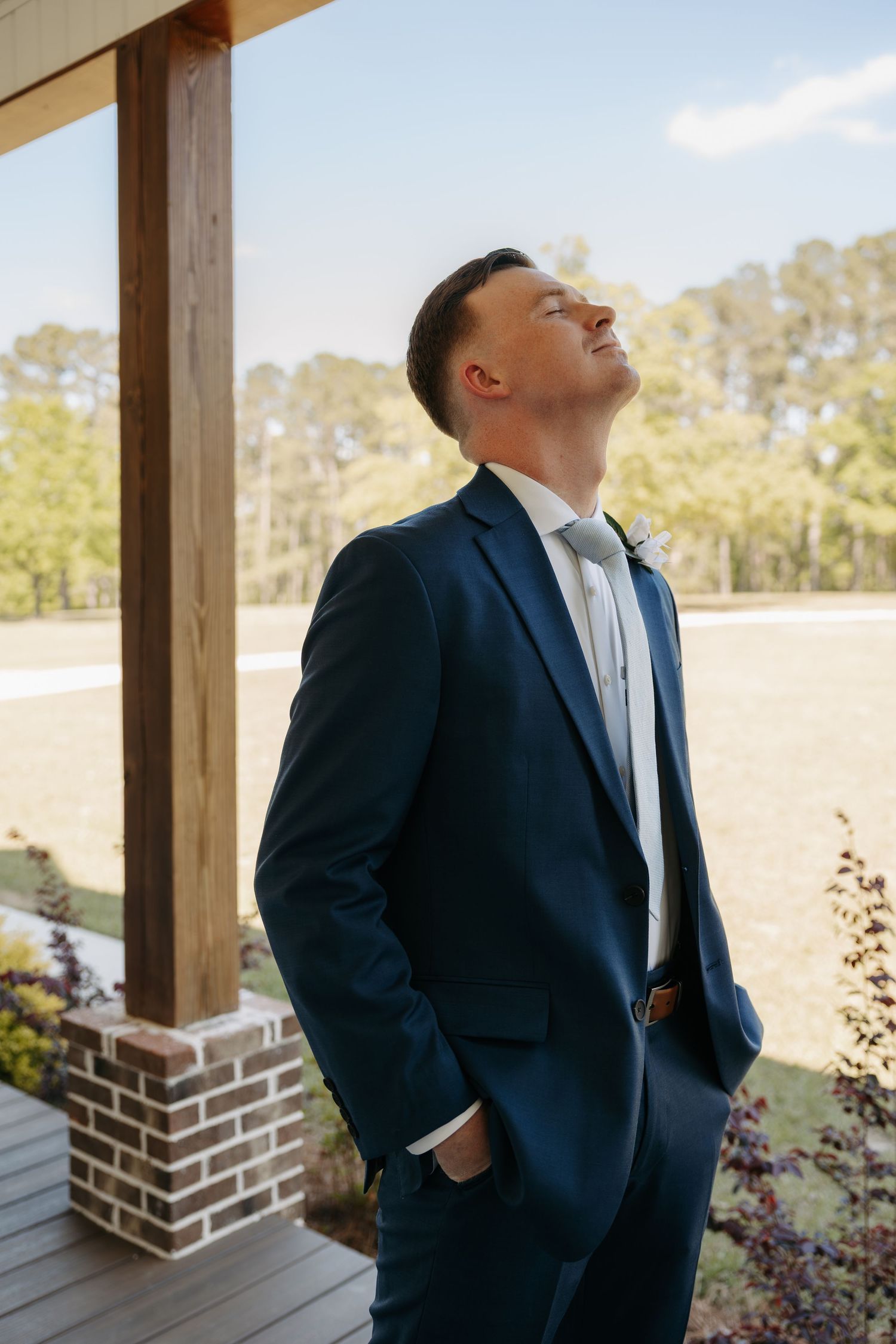 Groom in navy blue suit with white tie and boutonniere tilts head back on rustic porch at outdoor wedding venue.