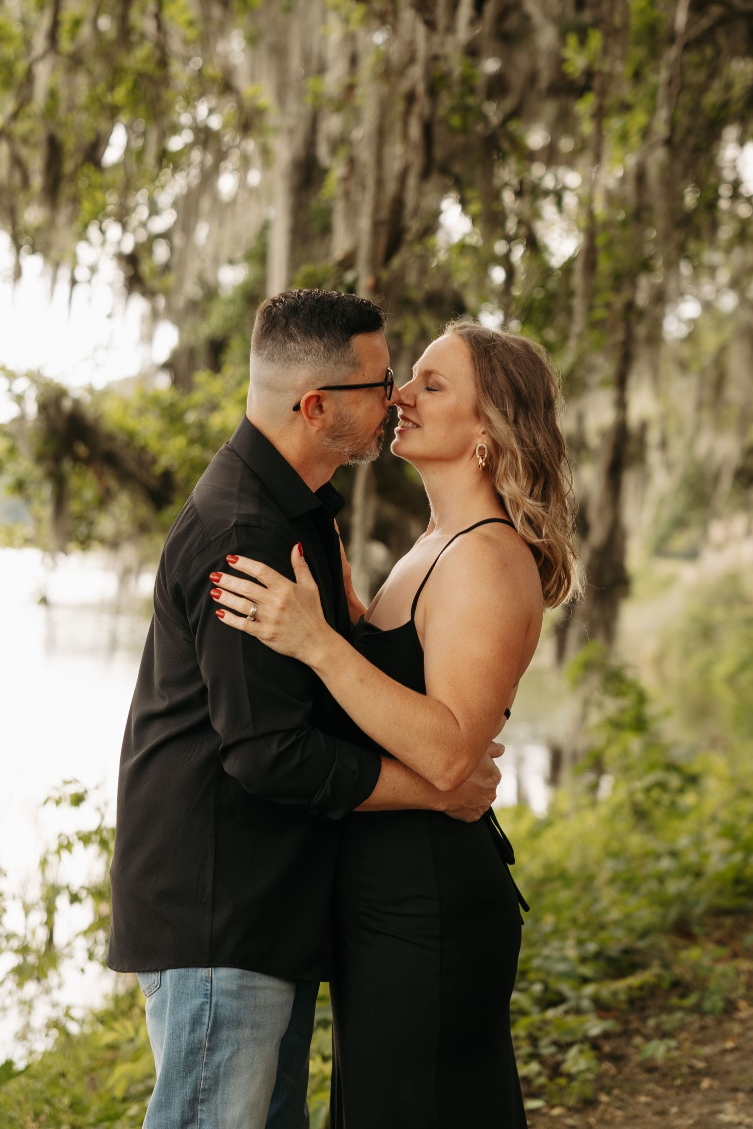 A couple in black outfits shares a romantic moment outdoors under moss-draped trees near a serene lake.