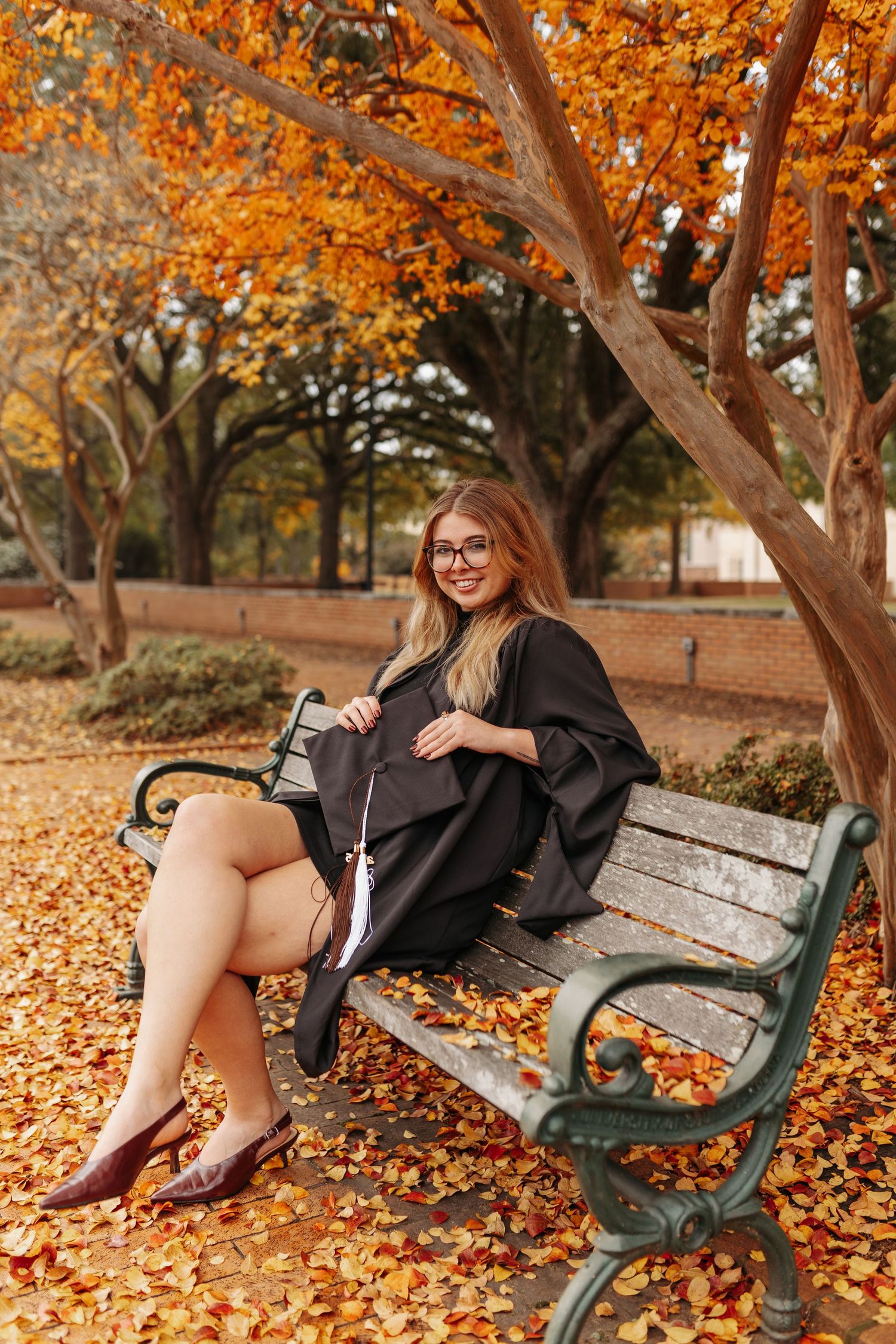 Graduate relaxes on park bench surrounded by vibrant autumn leaves and fall foliage.