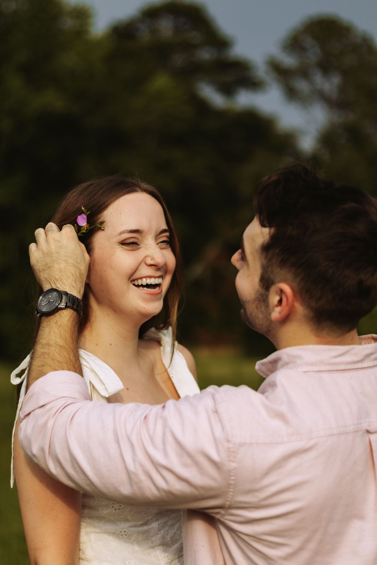 A laughing bride with flowers in her hair as her groom gently touches her face outdoors.