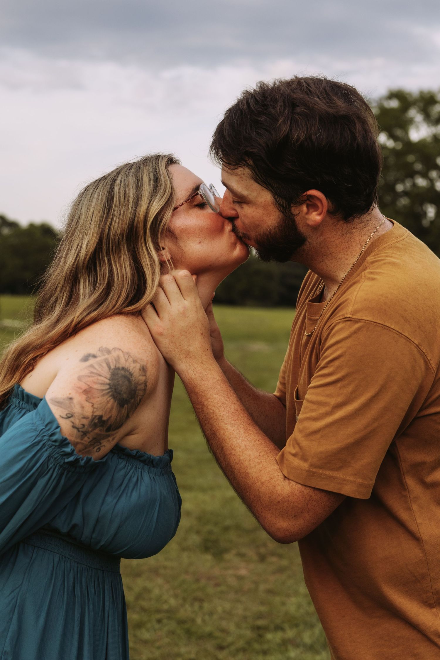 A couple shares a romantic kiss outdoors in a green field, woman in teal dress with floral tattoo, man in mustard shirt.
