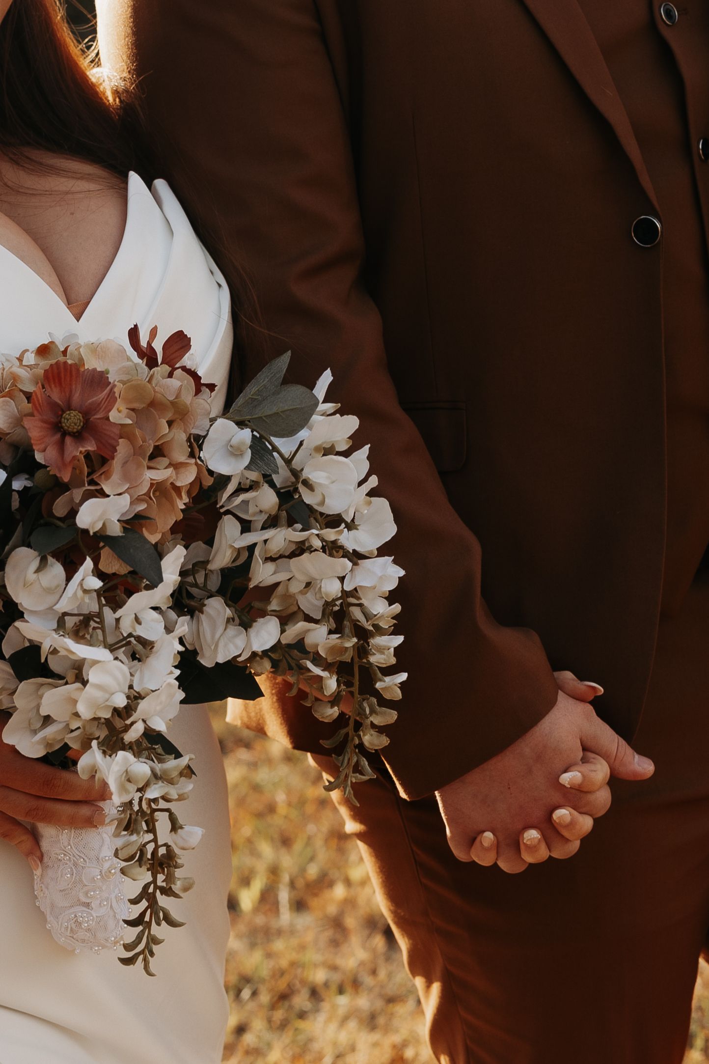 Bride and groom holding hands, wedding bouquet with white and dusty rose flowers, brown suit and white dress.