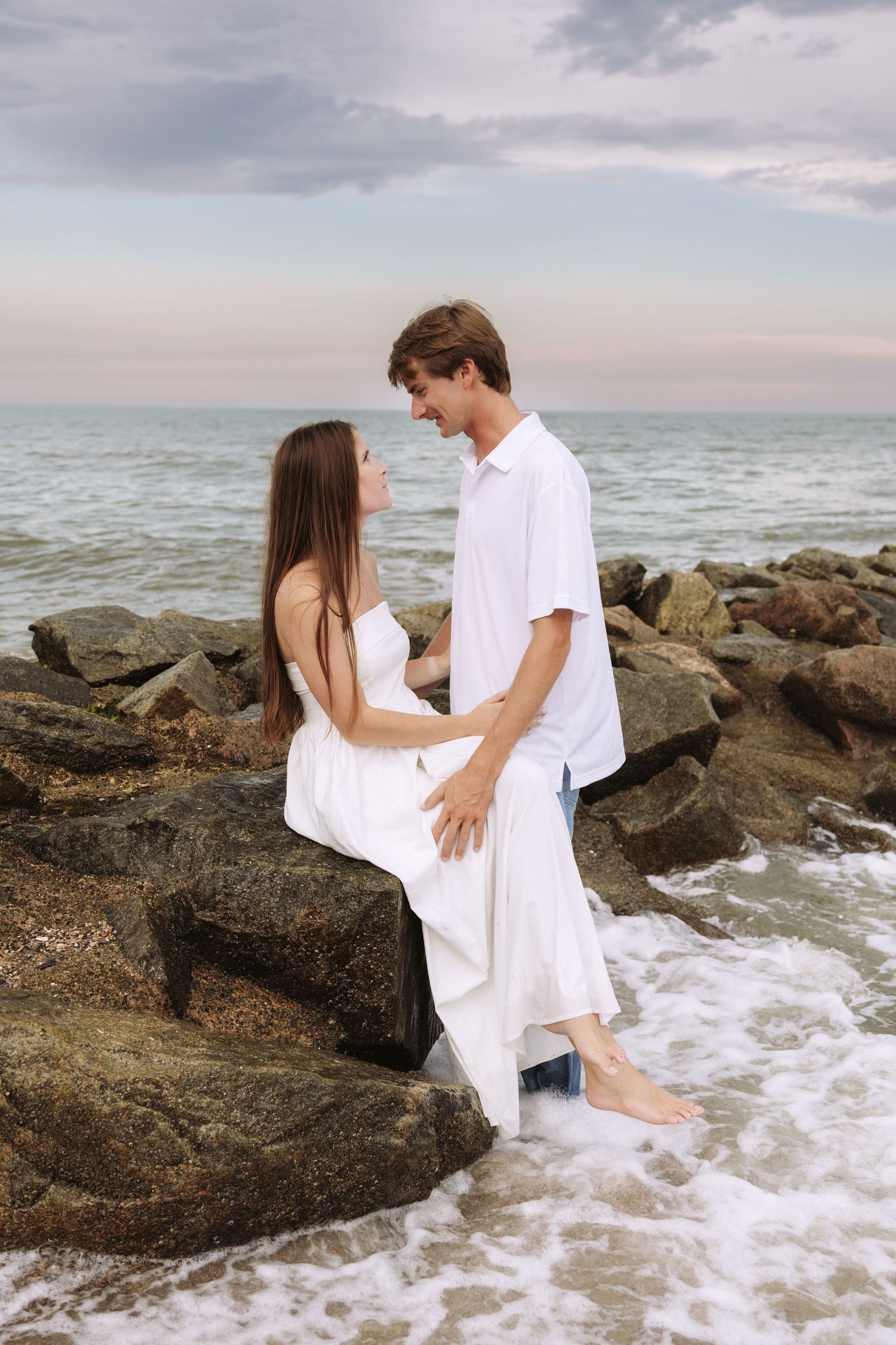 Romantic couple in white outfits sitting on rocky beach shore, gazing at each other with ocean waves in background.