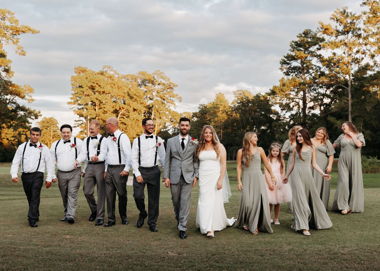 Wedding party walks across green lawn with groomsmen in gray suits with suspenders and bridesmaids in sage green gowns.