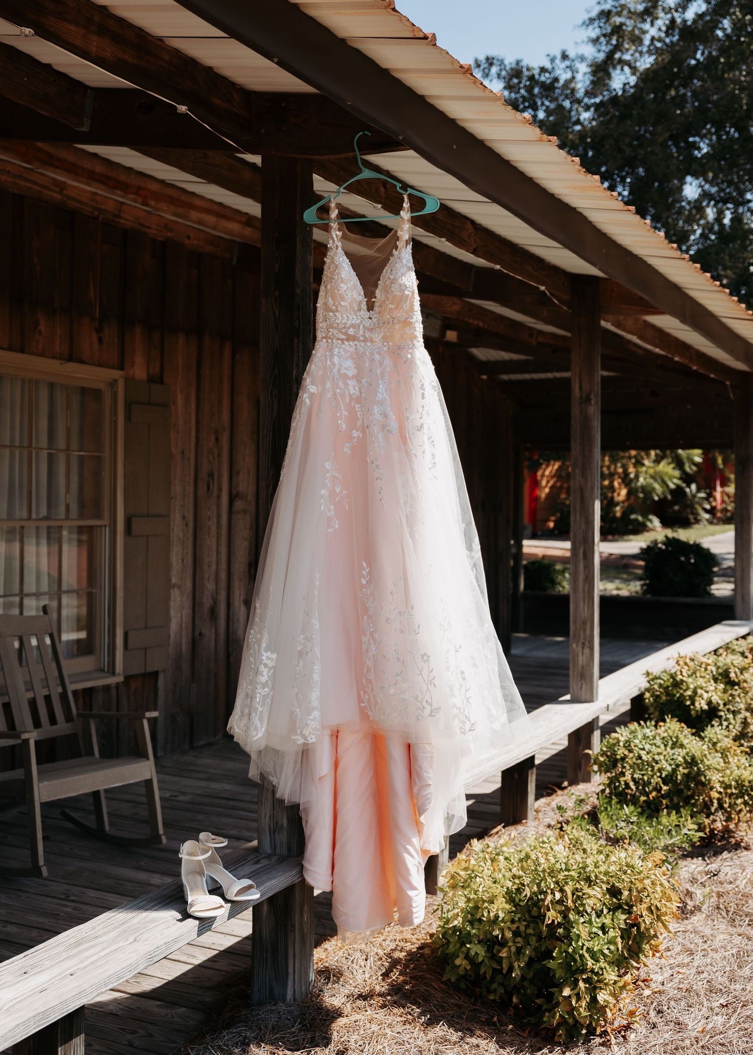 Blush lace wedding dress hanging on a rustic wooden porch of a countryside venue on a sunny day.