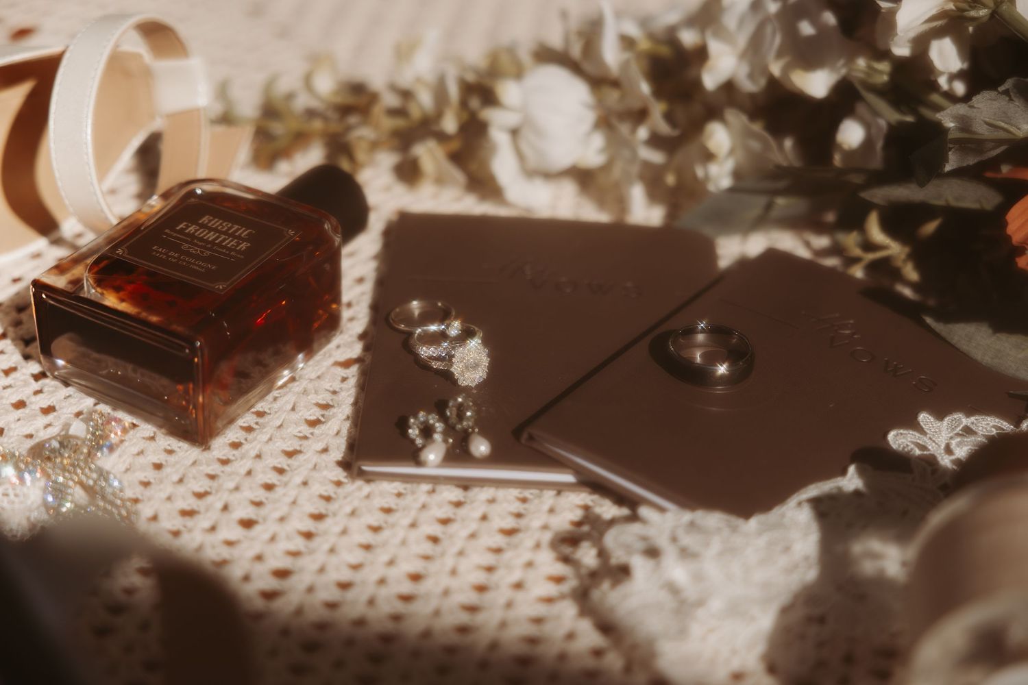 Elegant flat lay of amber perfume bottle, dark wedding stationery, diamond ring, and white florals in warm light.