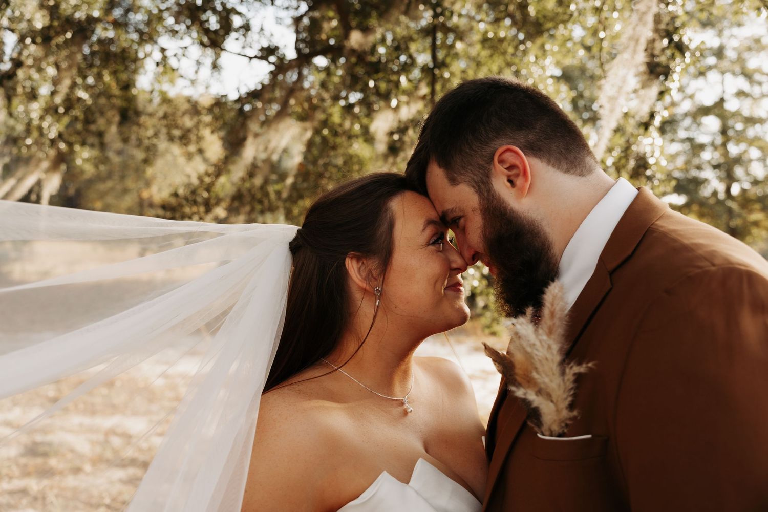 Bride and groom sharing a romantic forehead touch under a flowing veil outdoors on their wedding day.