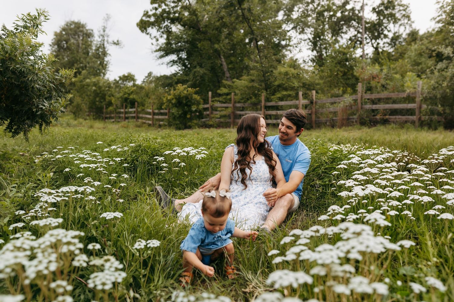 Family sitting in a wildflower field with white blooms, parents smiling at each other while child plays in grass.