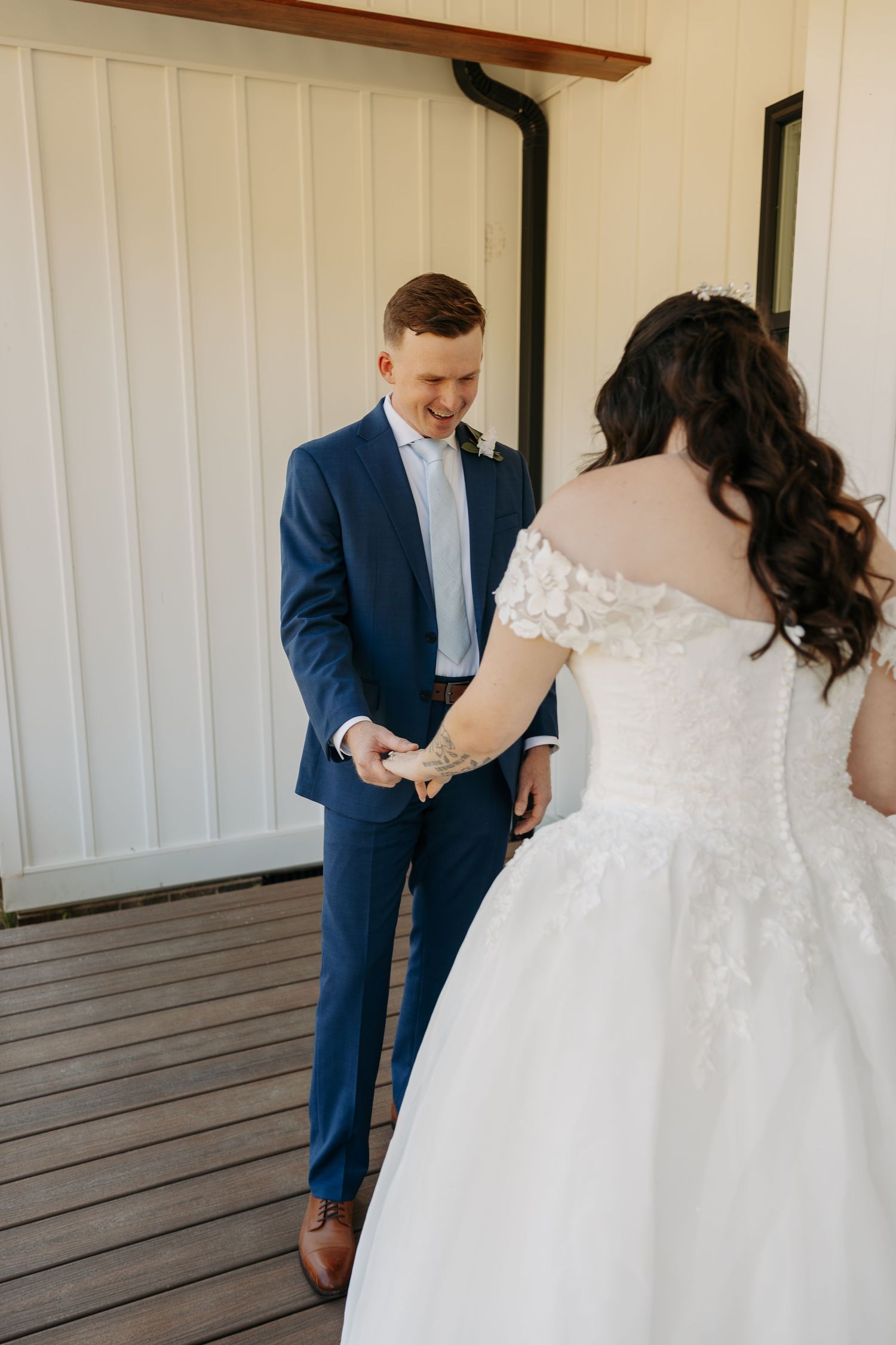Groom in navy suit smiling while holding bride's hand in white lace wedding gown on wooden porch.