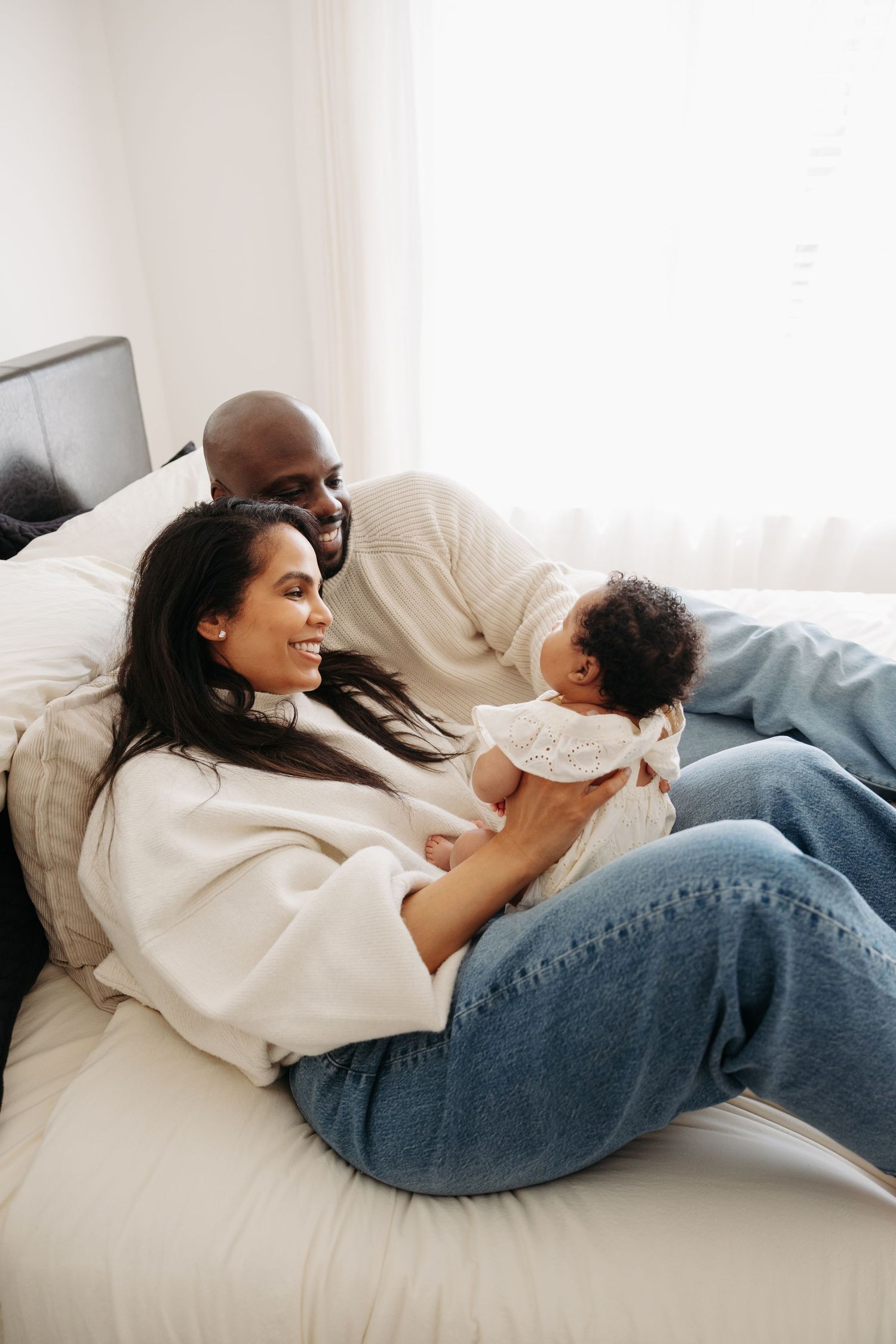 Happy family with newborn baby relaxing on bed, parents smiling and bonding with their infant at home.