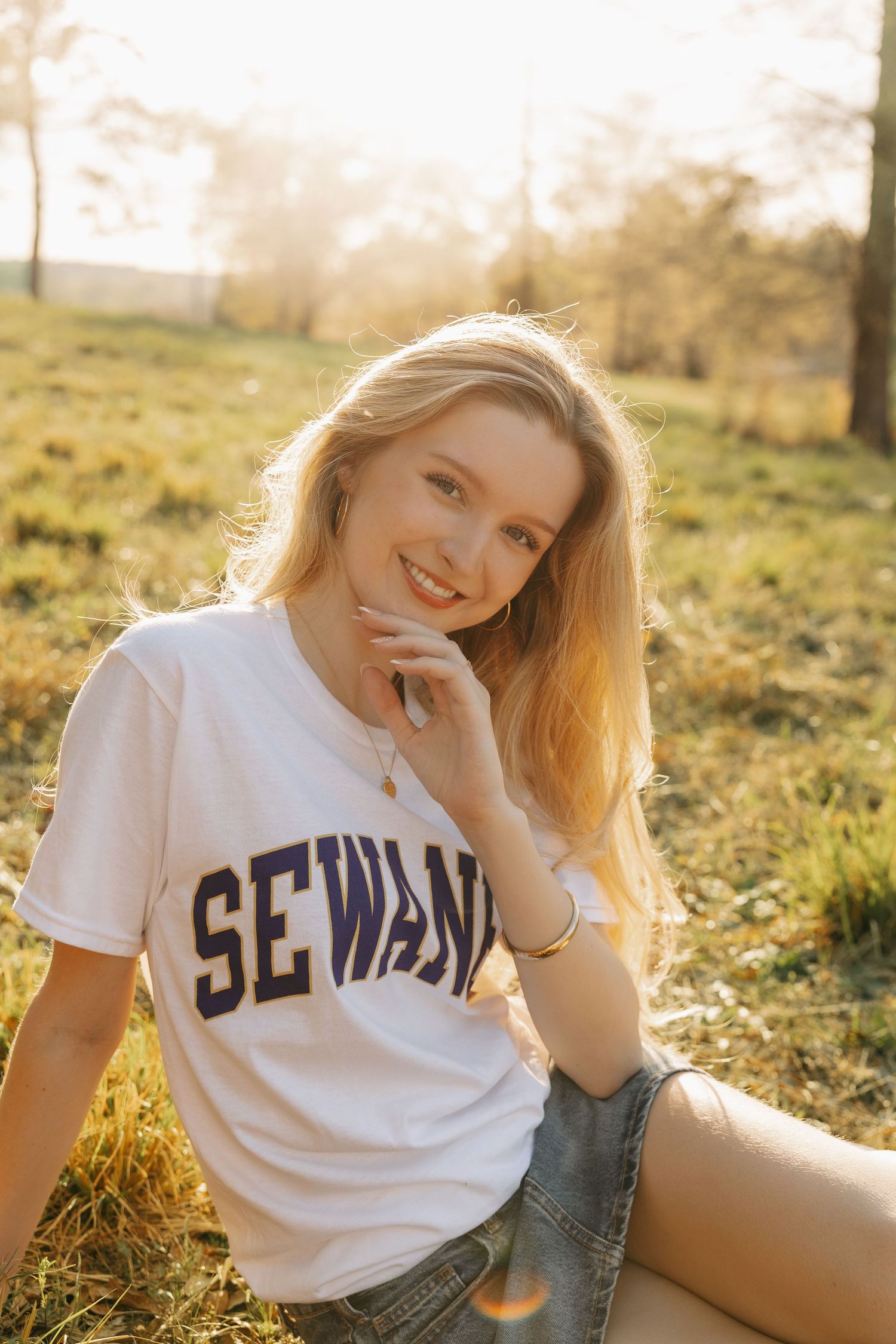 Smiling blonde woman in a white Sewanee t-shirt and denim shorts sitting on grass in golden sunlight.