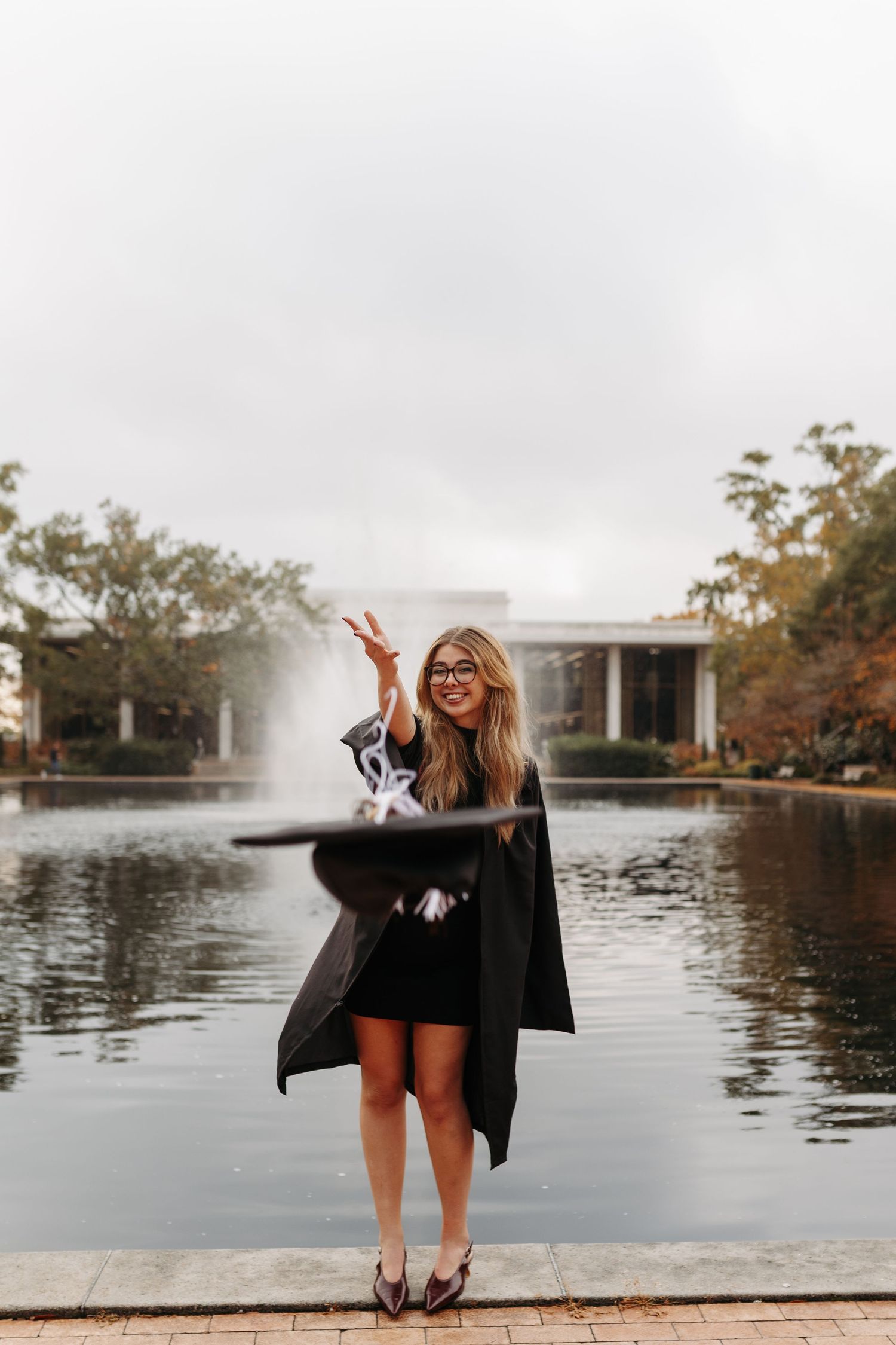 Graduate celebrating by a lake wearing a black graduation gown and shorts making peace signs at sunset.