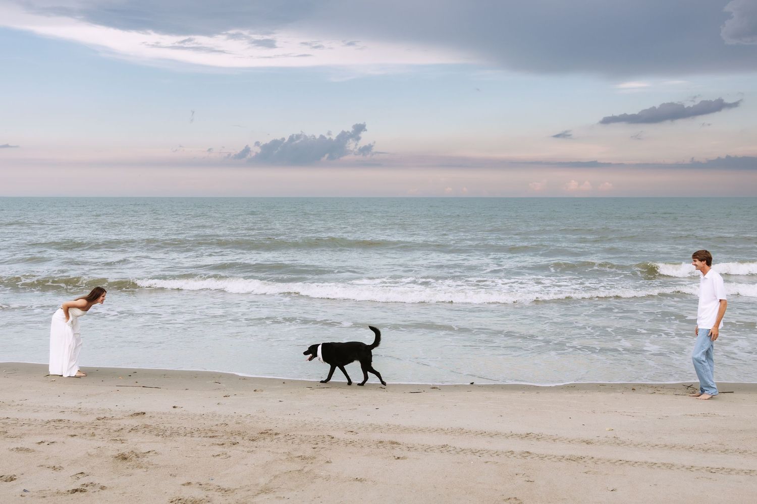 A couple enjoys a sunset beach walk with their black dog along the shoreline as gentle waves roll in.