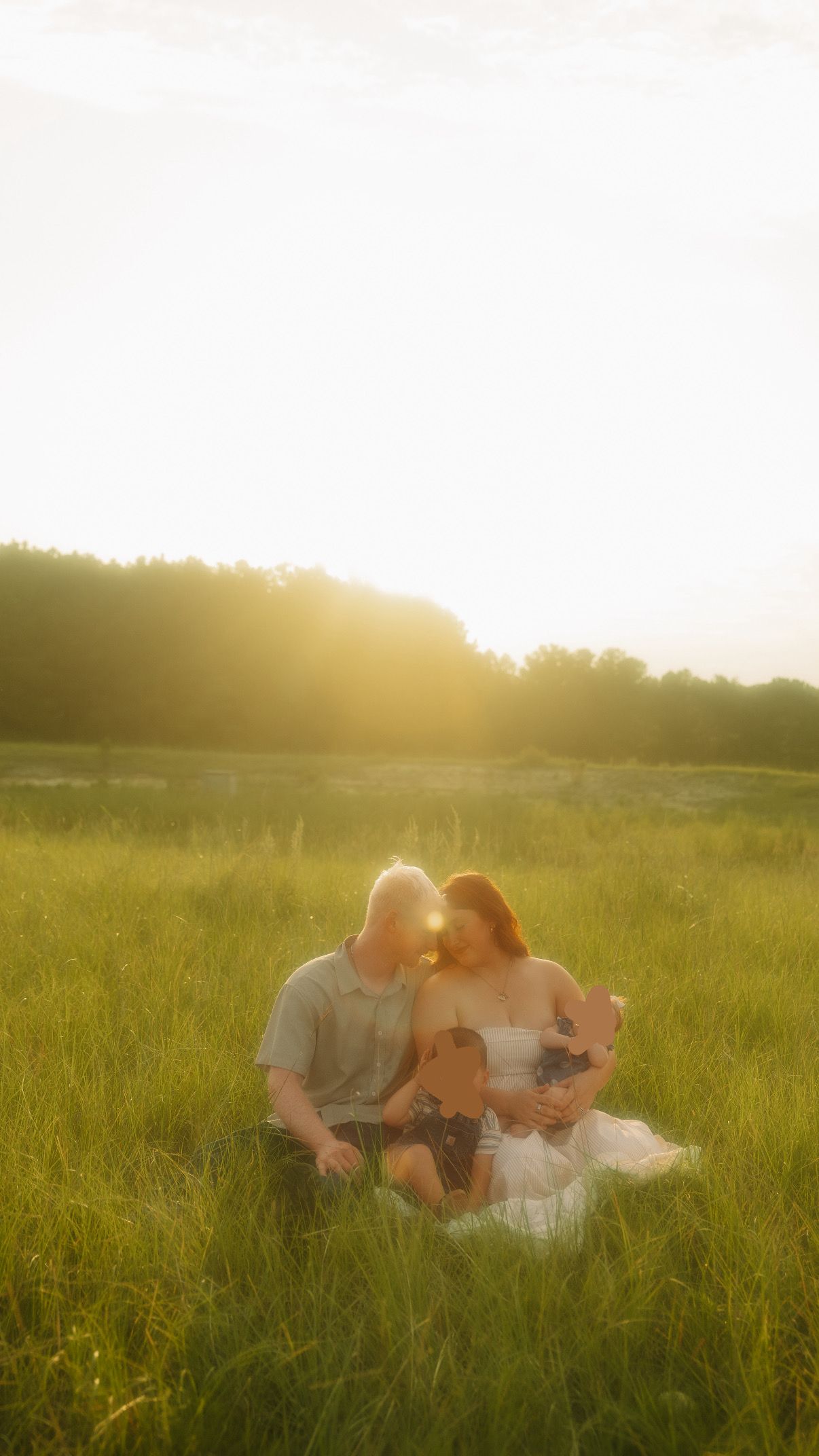 Family sitting together in a sunlit green meadow at golden hour, sharing a tender moment with warm glowing backlight.