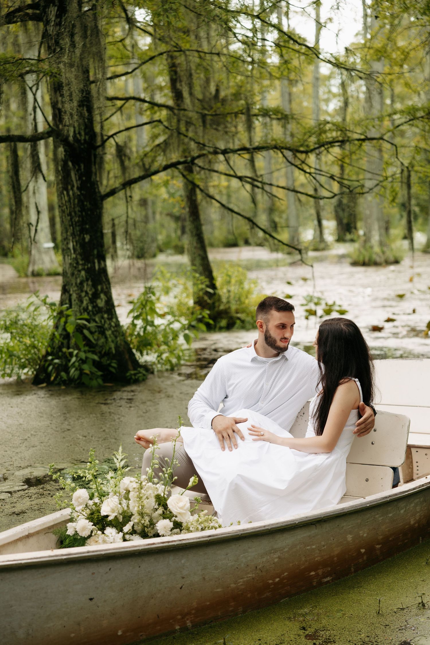 Couple in white attire sitting romantically in a boat with white flowers on a serene, tree-lined pond.
