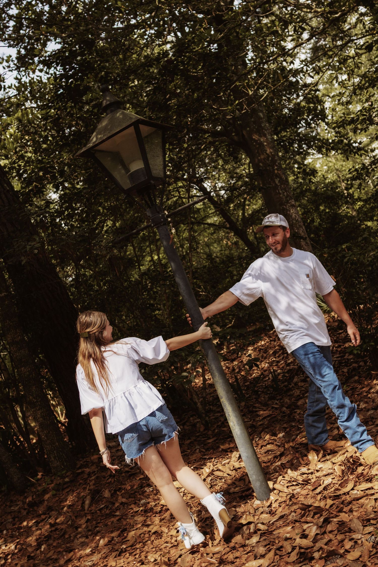 A couple holds hands around a lamp post in a sunlit forest, both wearing white tops and jeans.
