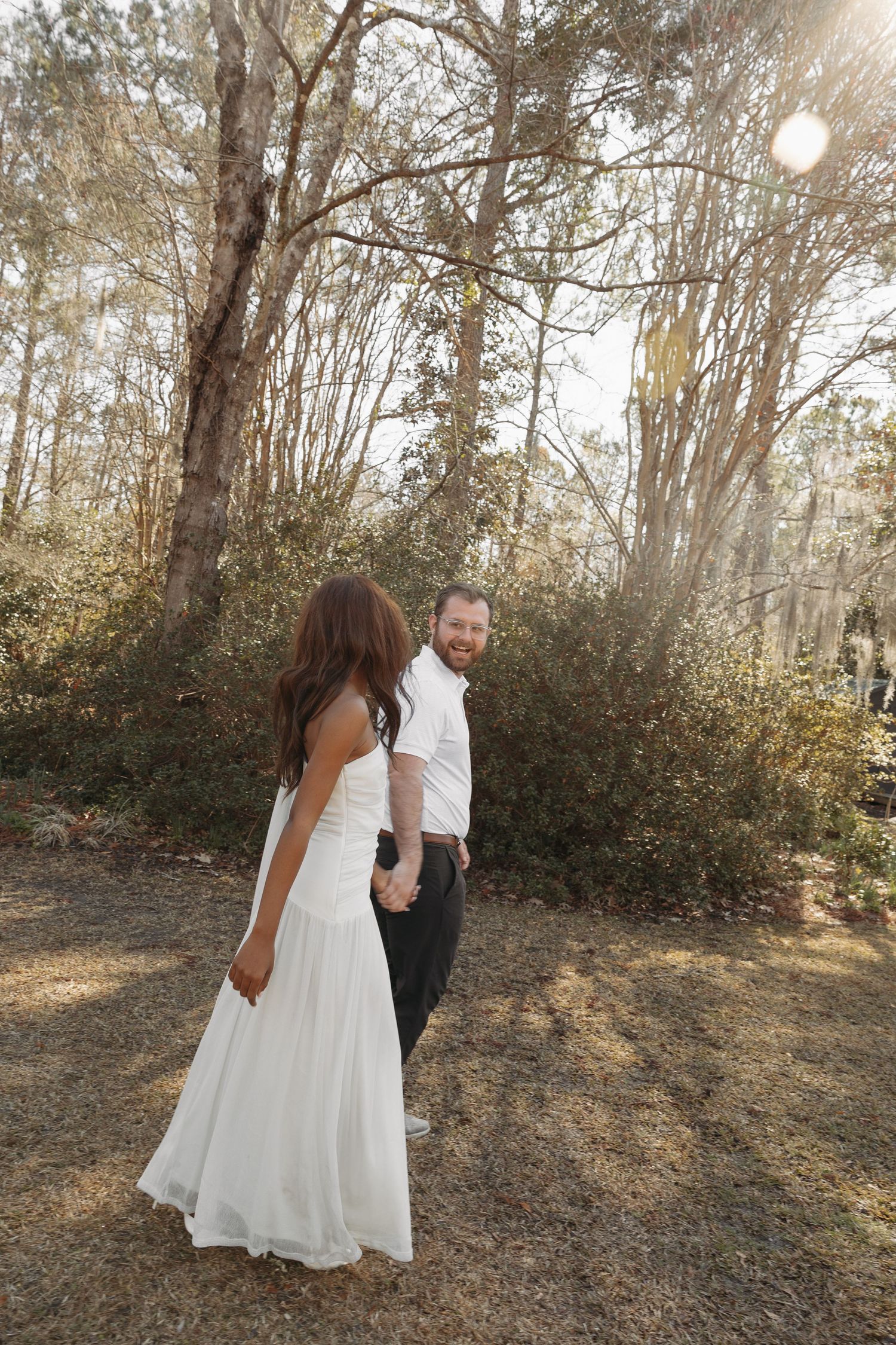 A bride in a white gown and groom in a white shirt hold hands while walking through a sunlit forest.