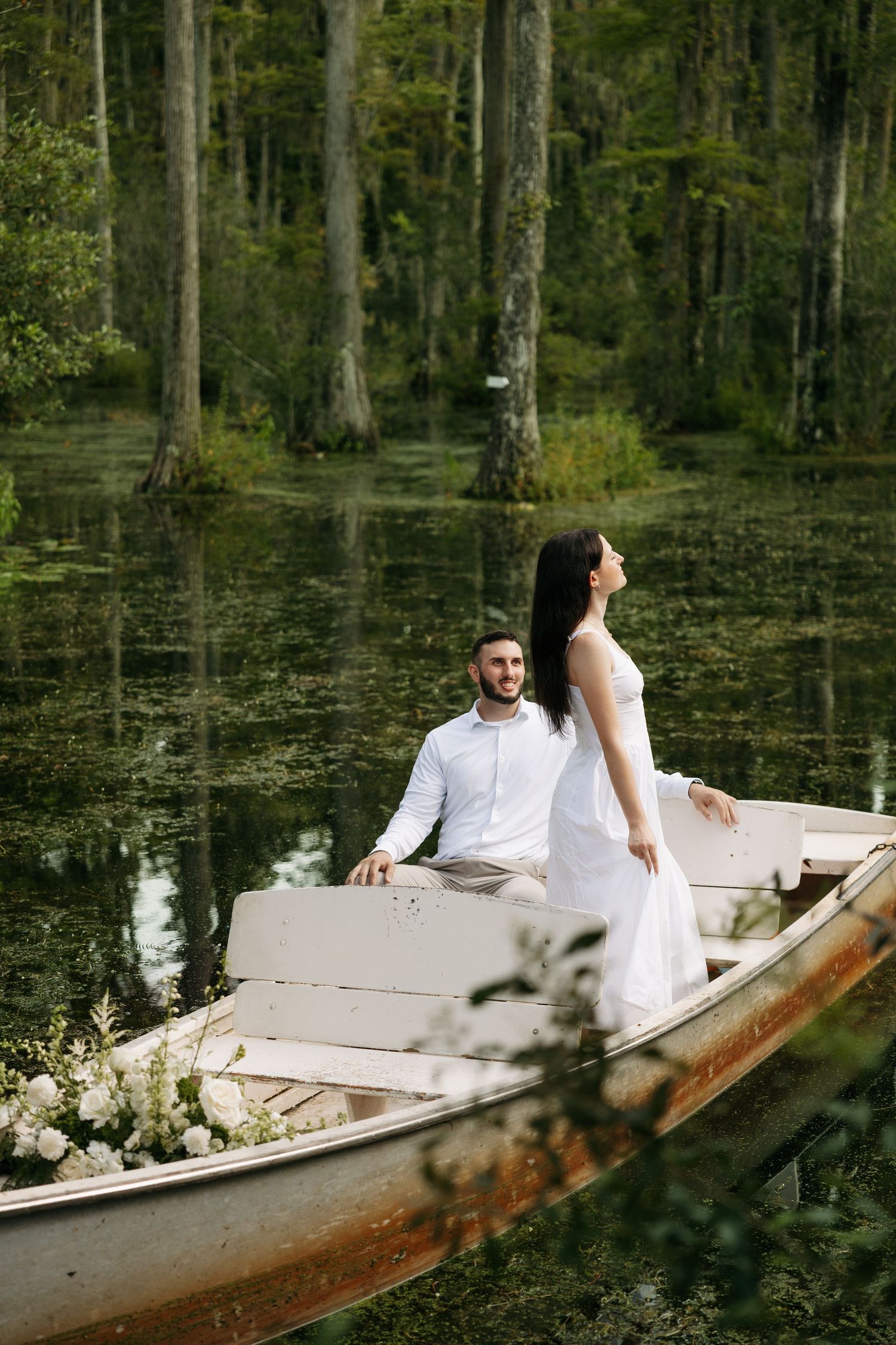Couple in white attire standing in a decorated rowboat surrounded by a lush swamp forest.