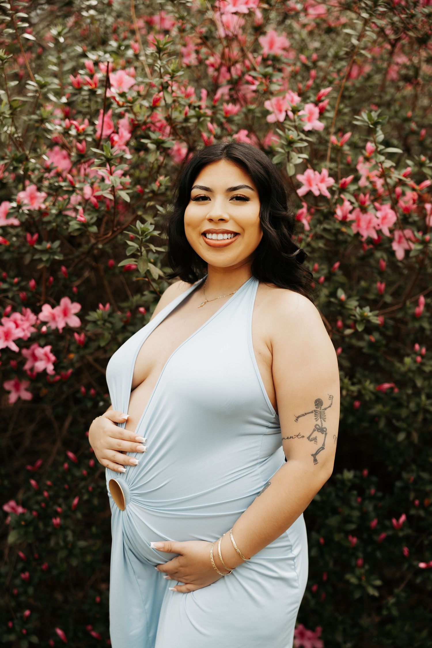 Smiling pregnant woman in light blue halter dress poses in front of pink flowering bushes, showing baby bump and arm tattoo.