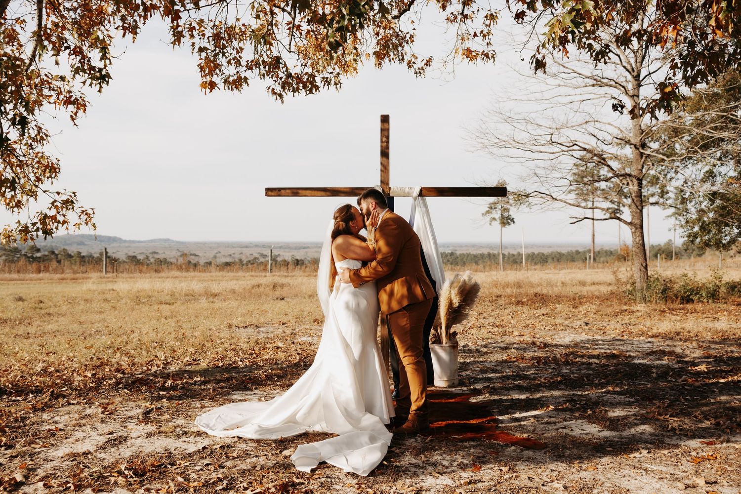 Bride and groom kissing before rustic wooden cross altar in autumn field at outdoor wedding ceremony.